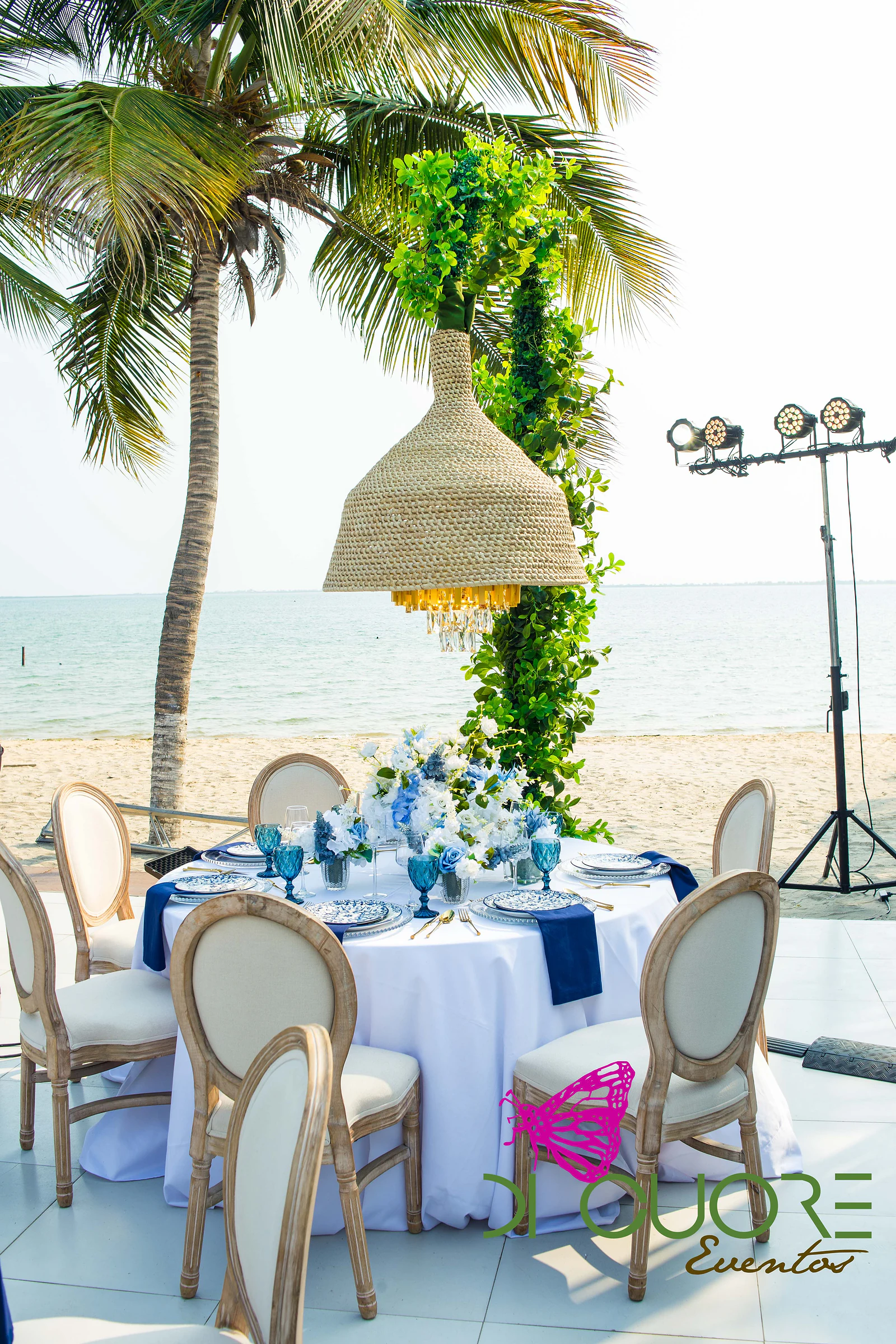 Round table in white linen with blue plates, white and blue flowers and a straw pendant lamp hanging from a leafy stem, beside the beach and the sea