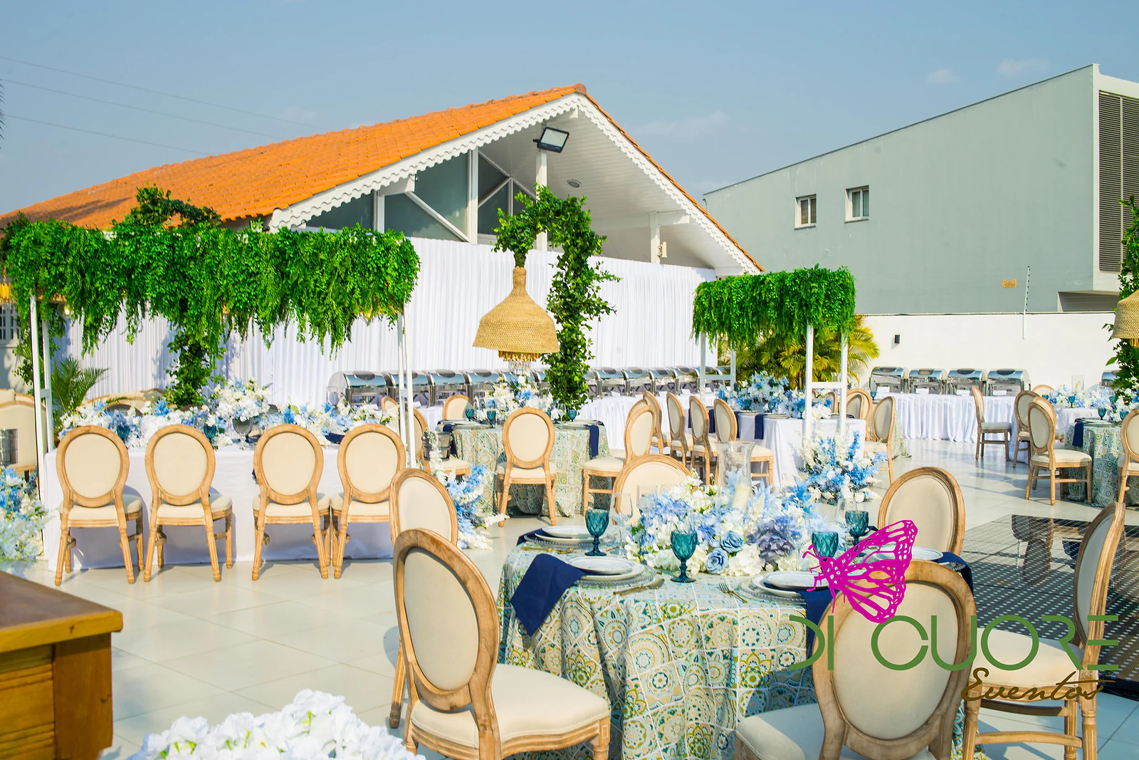 Round tables in patterned cloths with timber chairs, blue flowers, straw lamps and frames hung with foliage, in front of a tile-roofed house