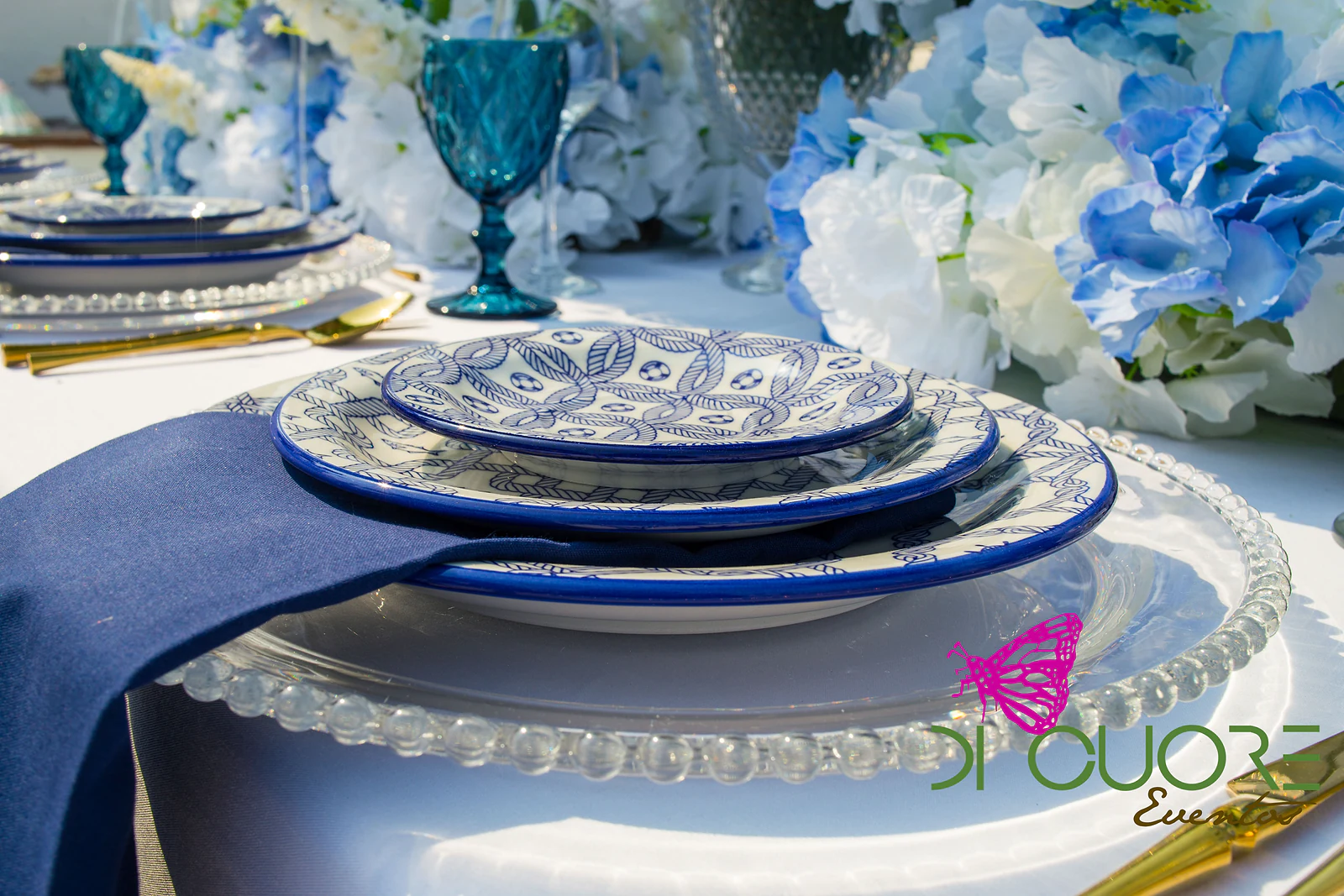Stack of blue-and-white plates on a beaded glass charger with a navy napkin, beside a teal glass and blue hydrangeas