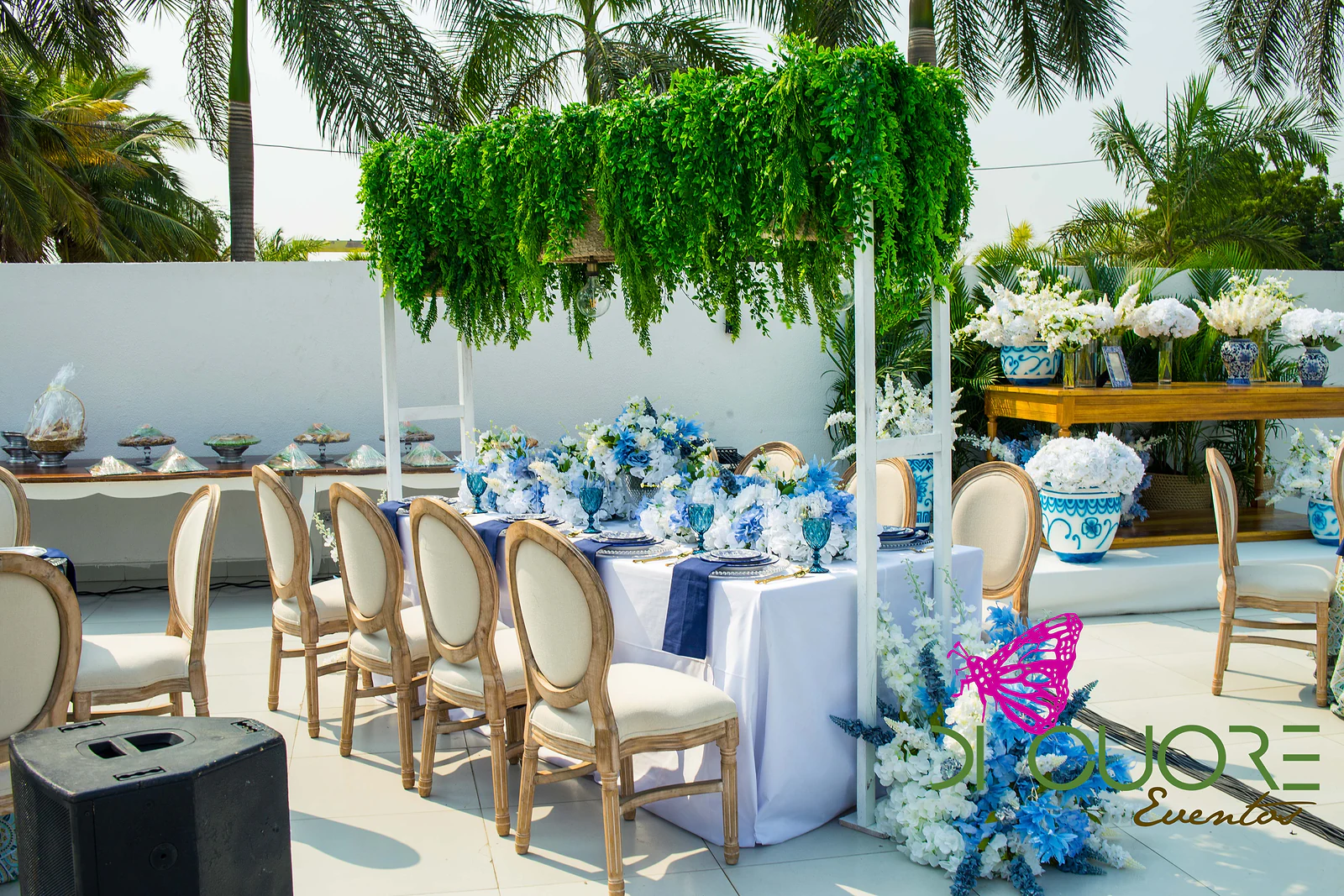 Long white-clothed table with navy napkins and blue flowers beneath a frame of hanging foliage, with the sideboard of blue-and-white vases beyond