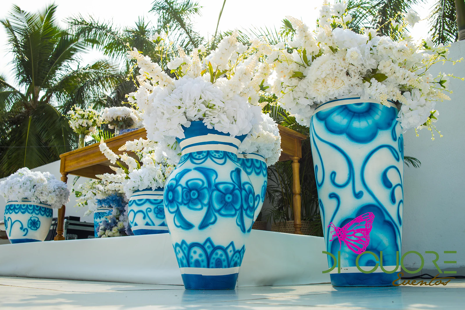 Tall blue-and-white painted vases holding sprays of white flowers on a timber sideboard, with palms beyond