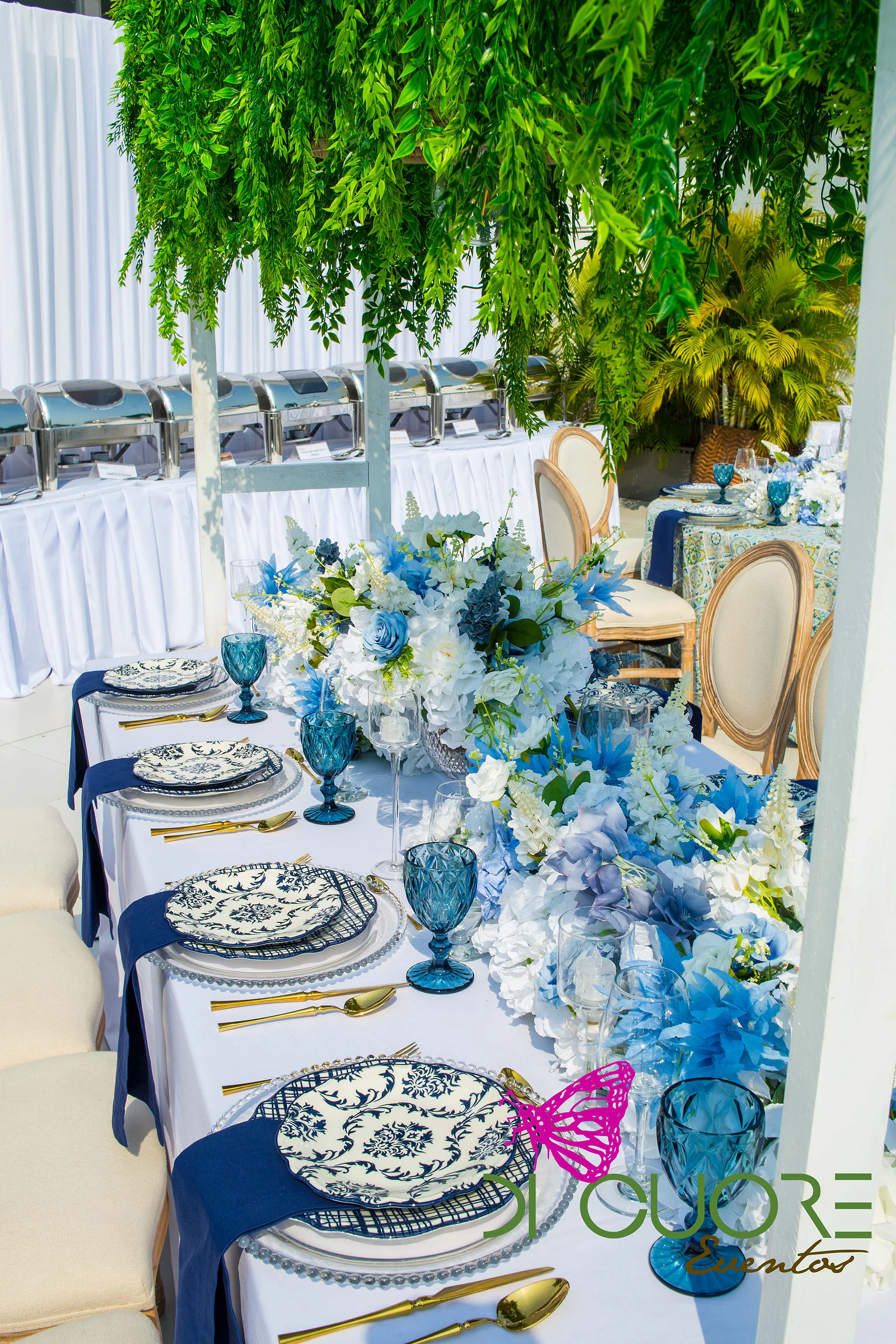 Long table with blue-and-white plates, navy napkins, teal glasses, gold cutlery and a large arrangement of blue and white hydrangeas beneath cascades of foliage