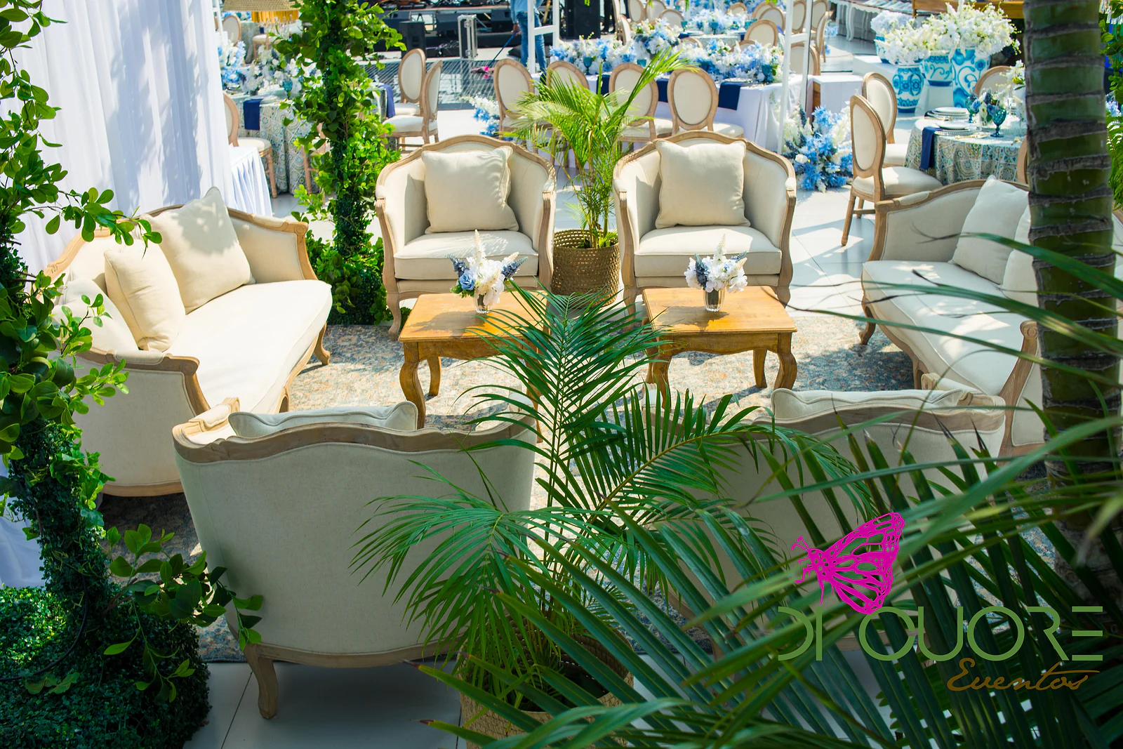 Timber-framed sofas and armchairs in cream upholstery around coffee tables, among palms and climbers, with laid tables beyond
