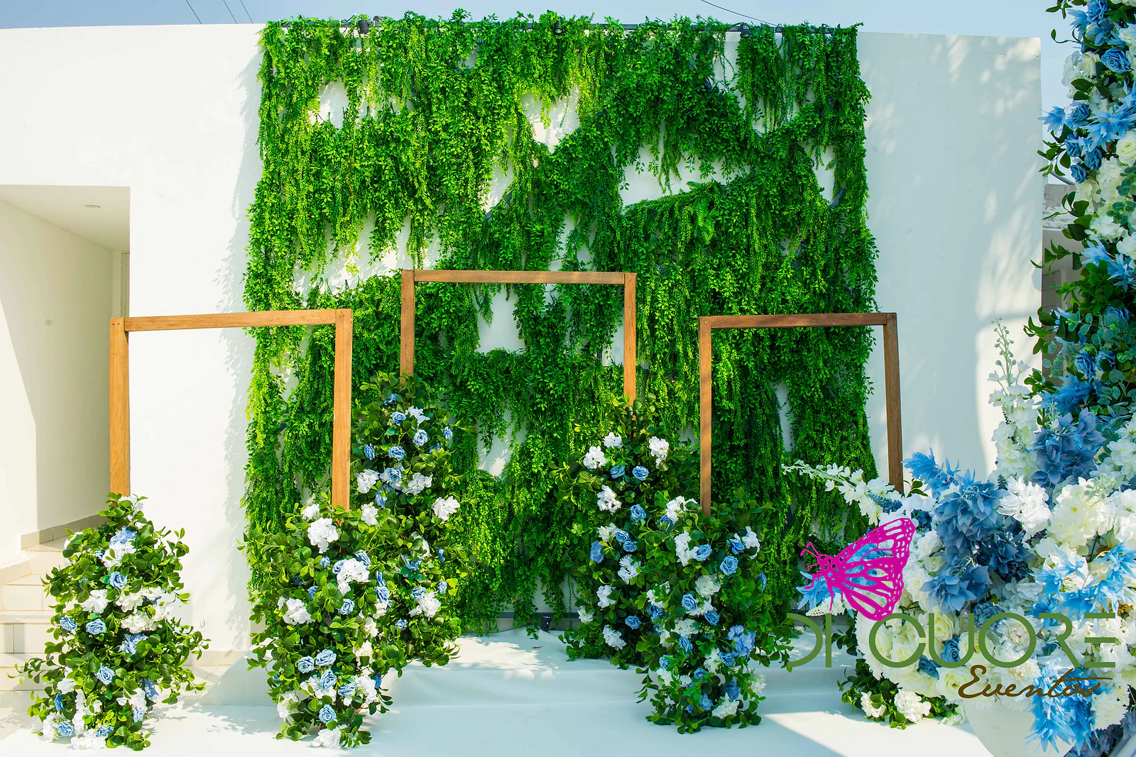 Wall of foliage with three timber frames and arrangements of blue and white flowers at its foot, on a white floor