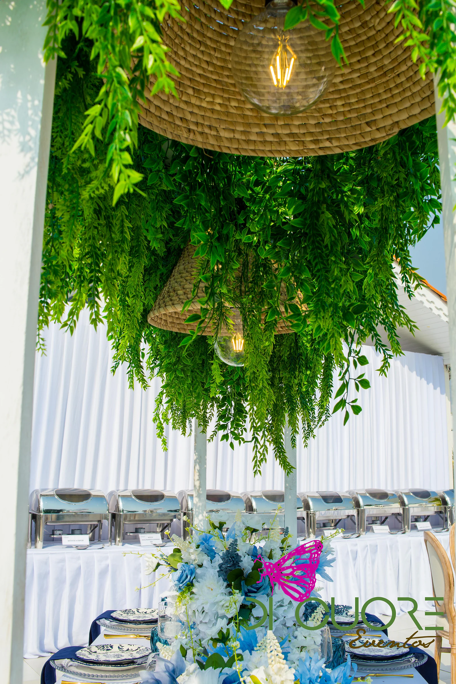 Straw pendant lamps with filament bulbs wrapped in cascades of foliage above a table of blue and white flowers, with buffet dishes beyond