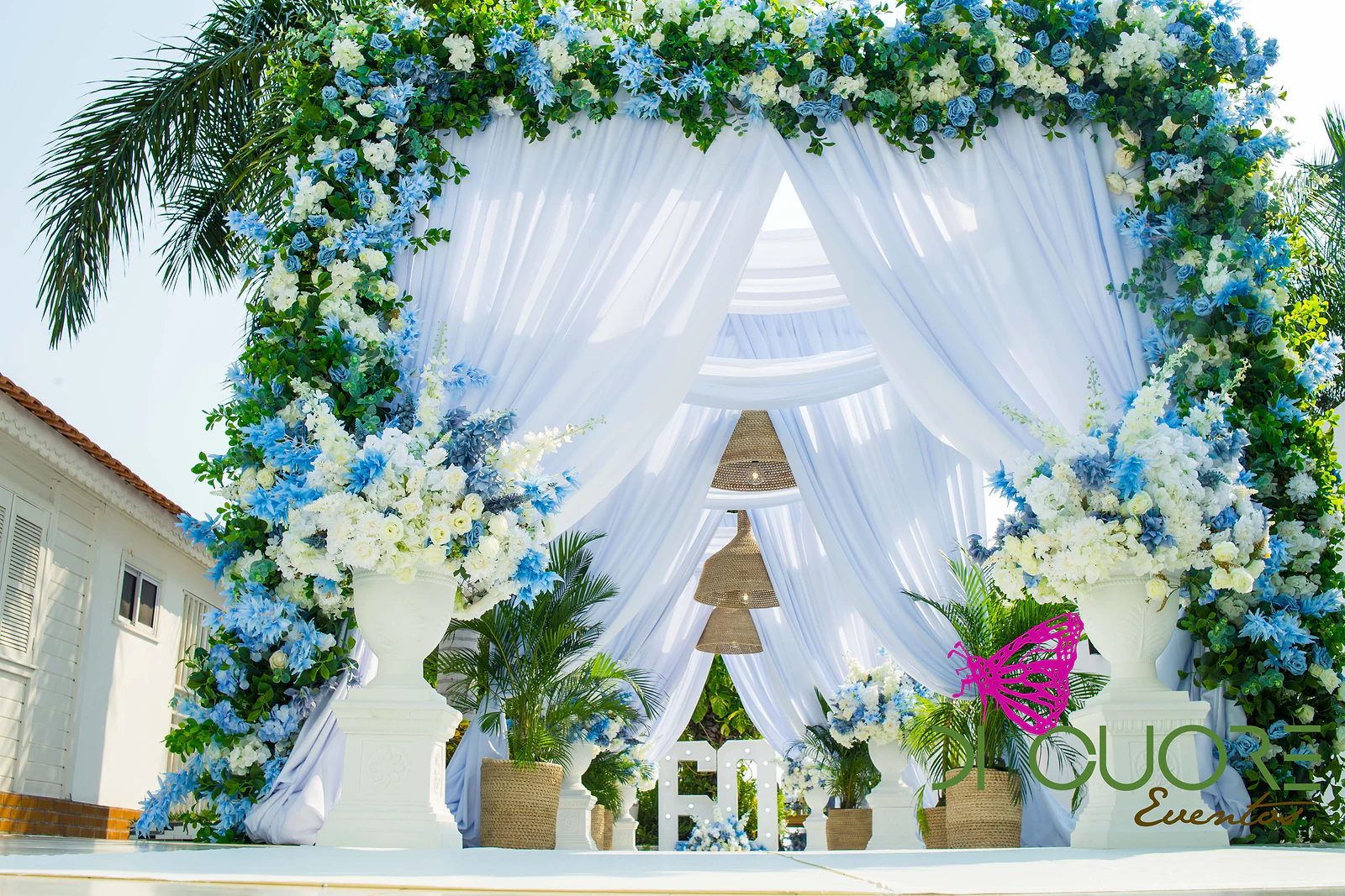 Arco de flores azuis e brancas com folhagem verde e cortinados brancos esvoaçantes, com candeeiros de vime e vasos com plantas à frente