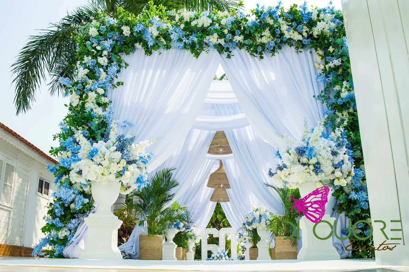 Gateway of blue and white flowers and foliage with white drapes, flowering urns on pedestals, palms in straw baskets and a lit-up number 60 beyond