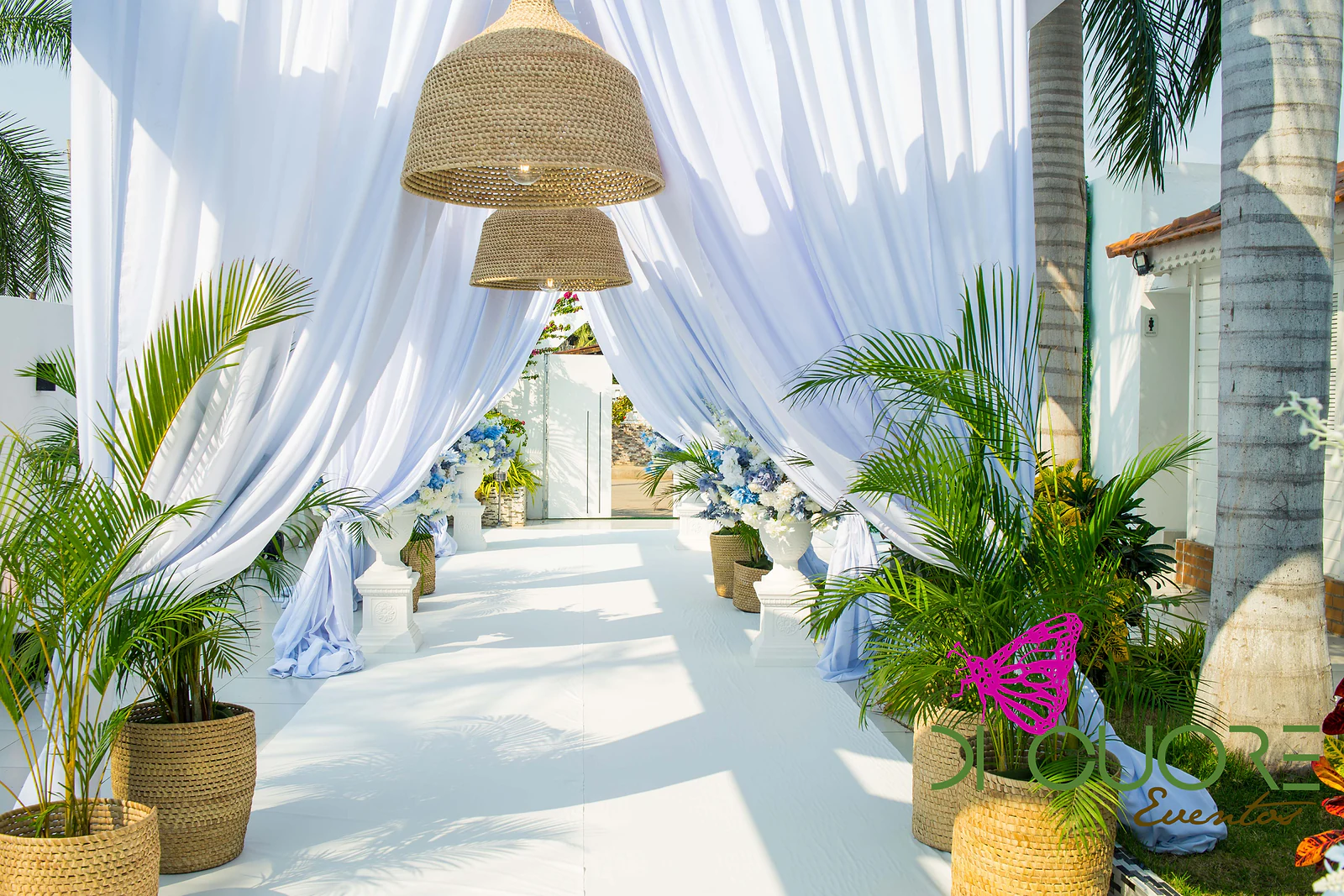 White walkway between billowing drapes, palms in straw baskets and hanging straw lamps