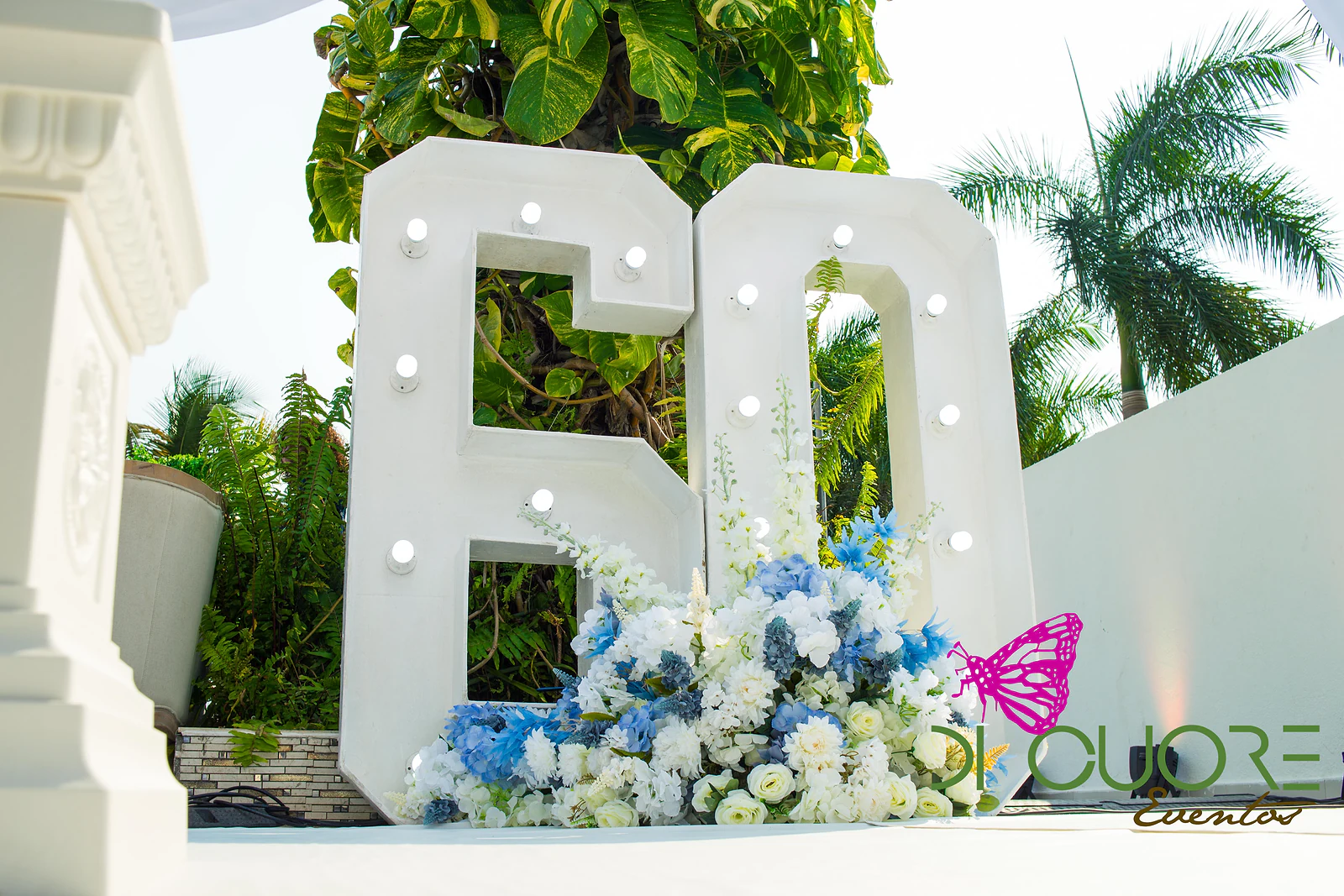 Large white number 60 in bulb-lit letters, with a generous bouquet of blue and white flowers at its foot and tropical foliage behind