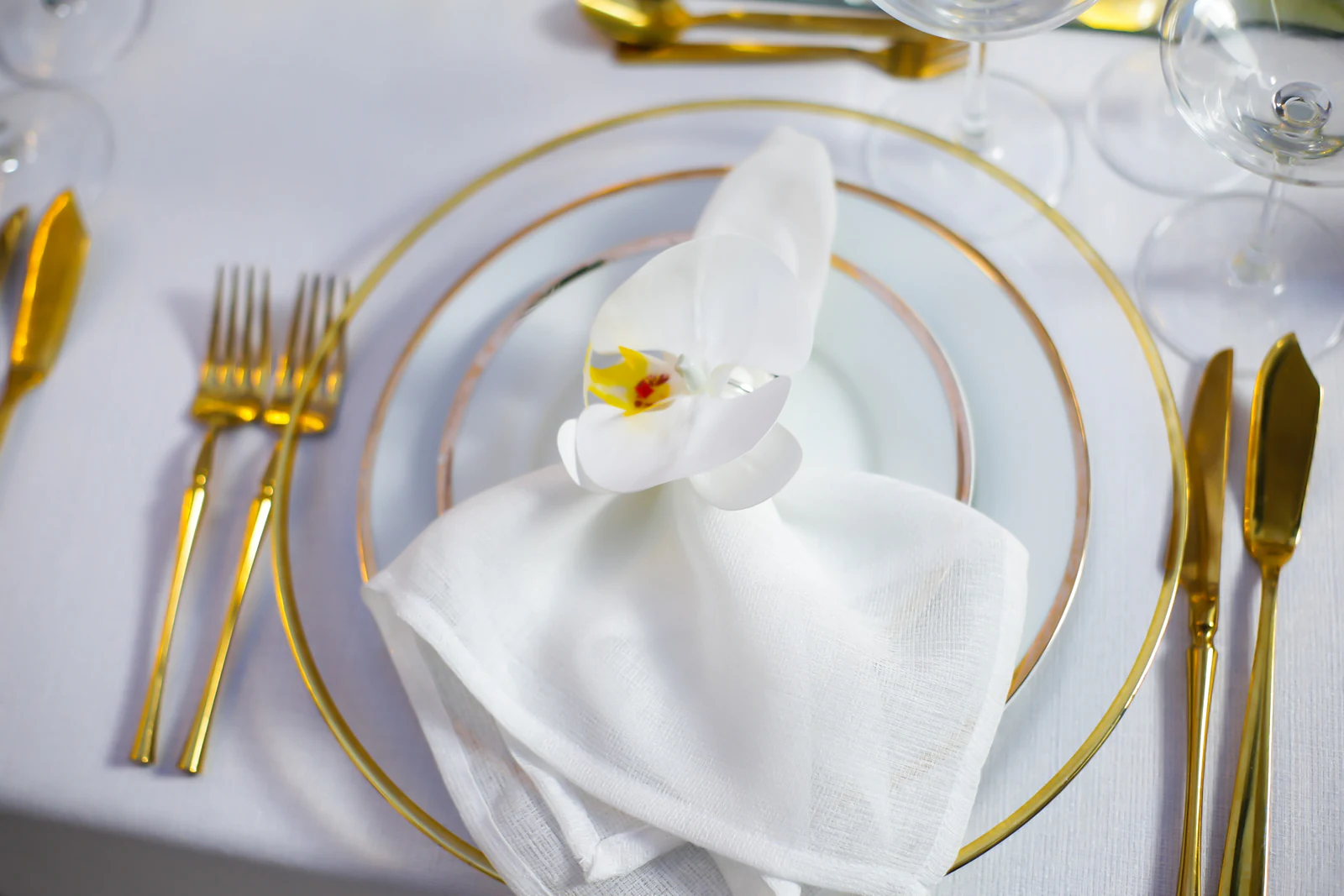 Prato de rebordo dourado com guardanapo de linho branco e uma orquídea, entre talheres dourados