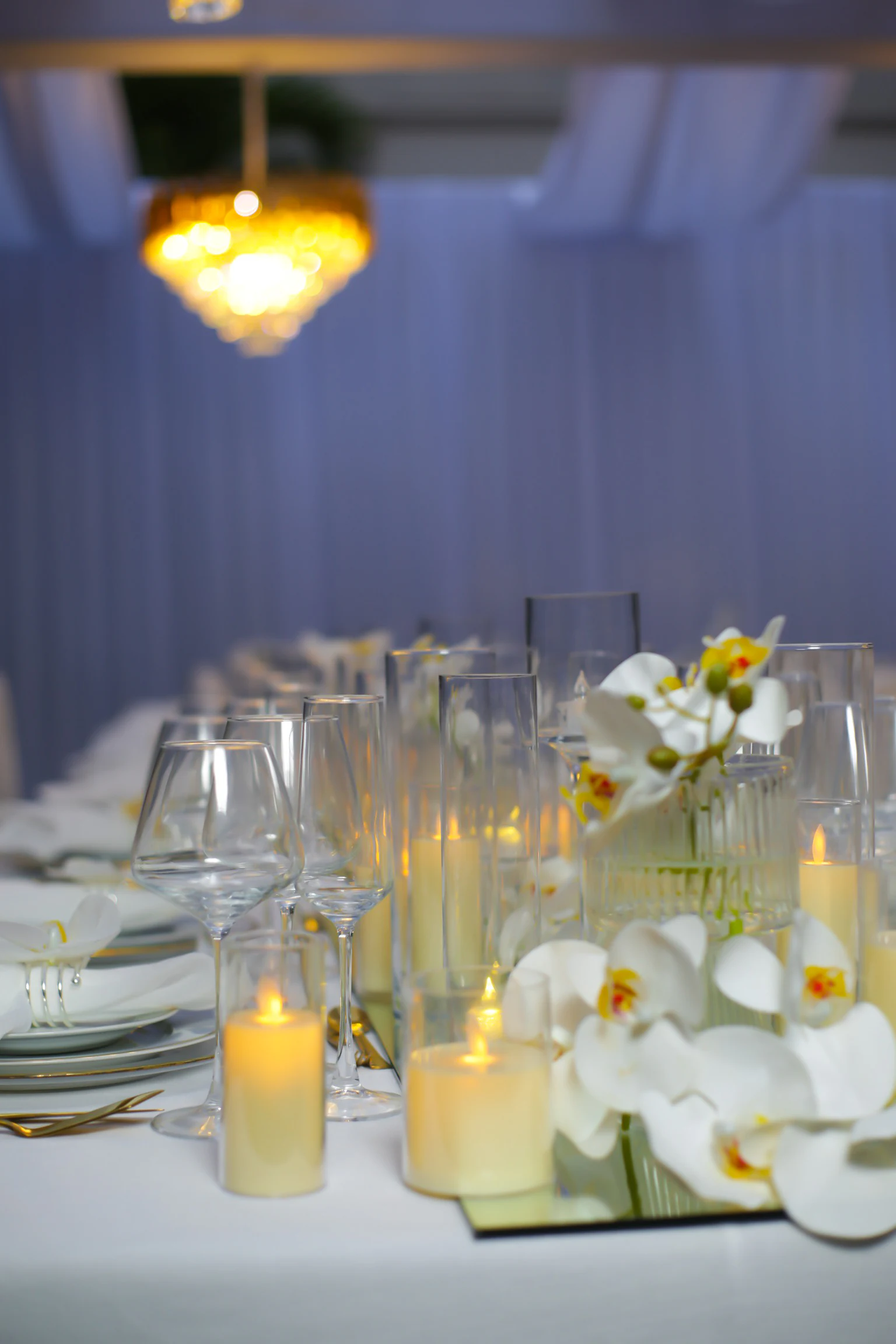 Orquídeas brancas e velas em copos de vidro sobre toalha branca, sob um lustre dourado e cortinados azulados