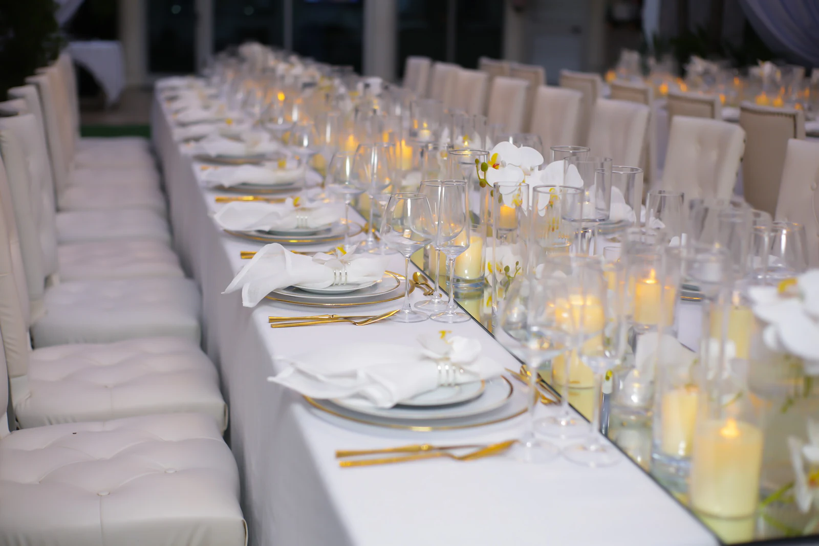 Mesa corrida vista de lado, com bancos estofados a branco, pratos postos e velas ao longo do friso de espelho