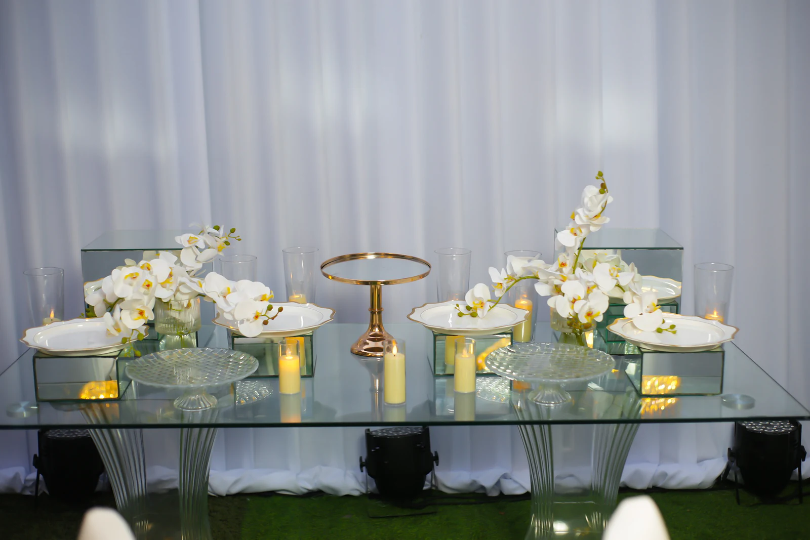 Mesa de vidro com cubos de espelho, pratos brancos, orquídeas, velas e um pé de bolo dourado, diante de um cortinado branco