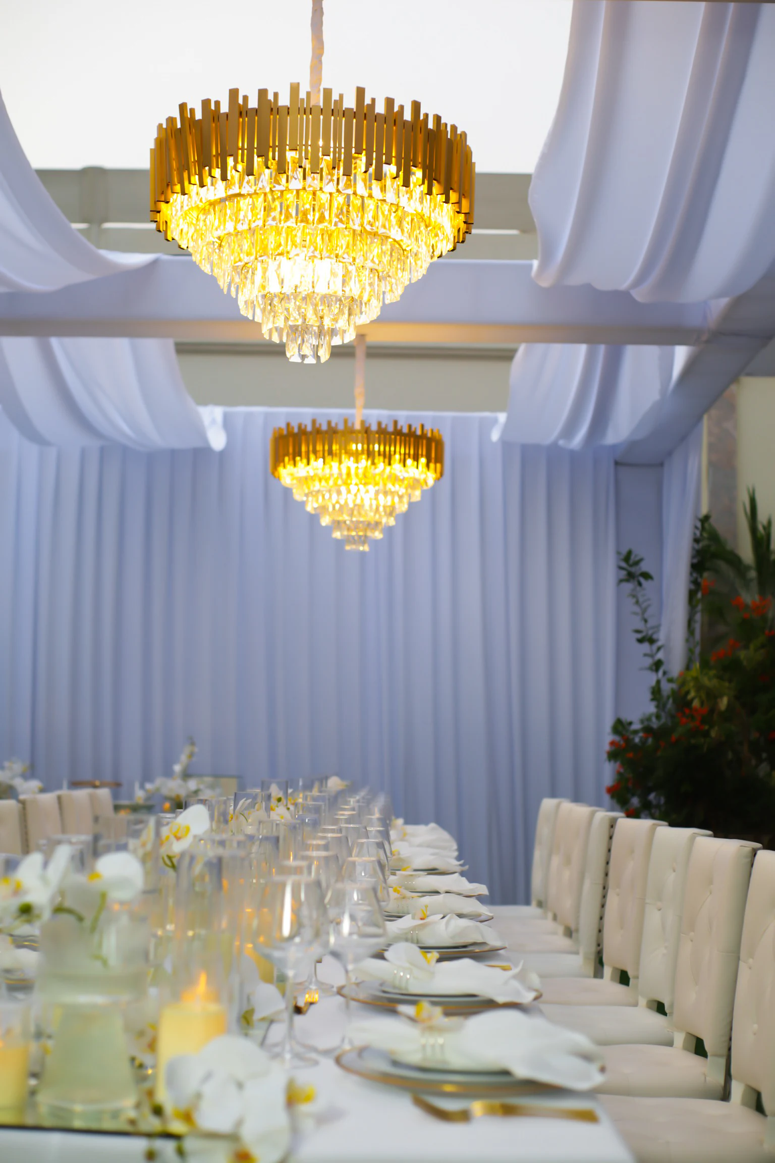 Dois lustres de cristal e latão dourado sobre a mesa posta, com guardanapos brancos, pratos de rebordo dourado e orquídeas