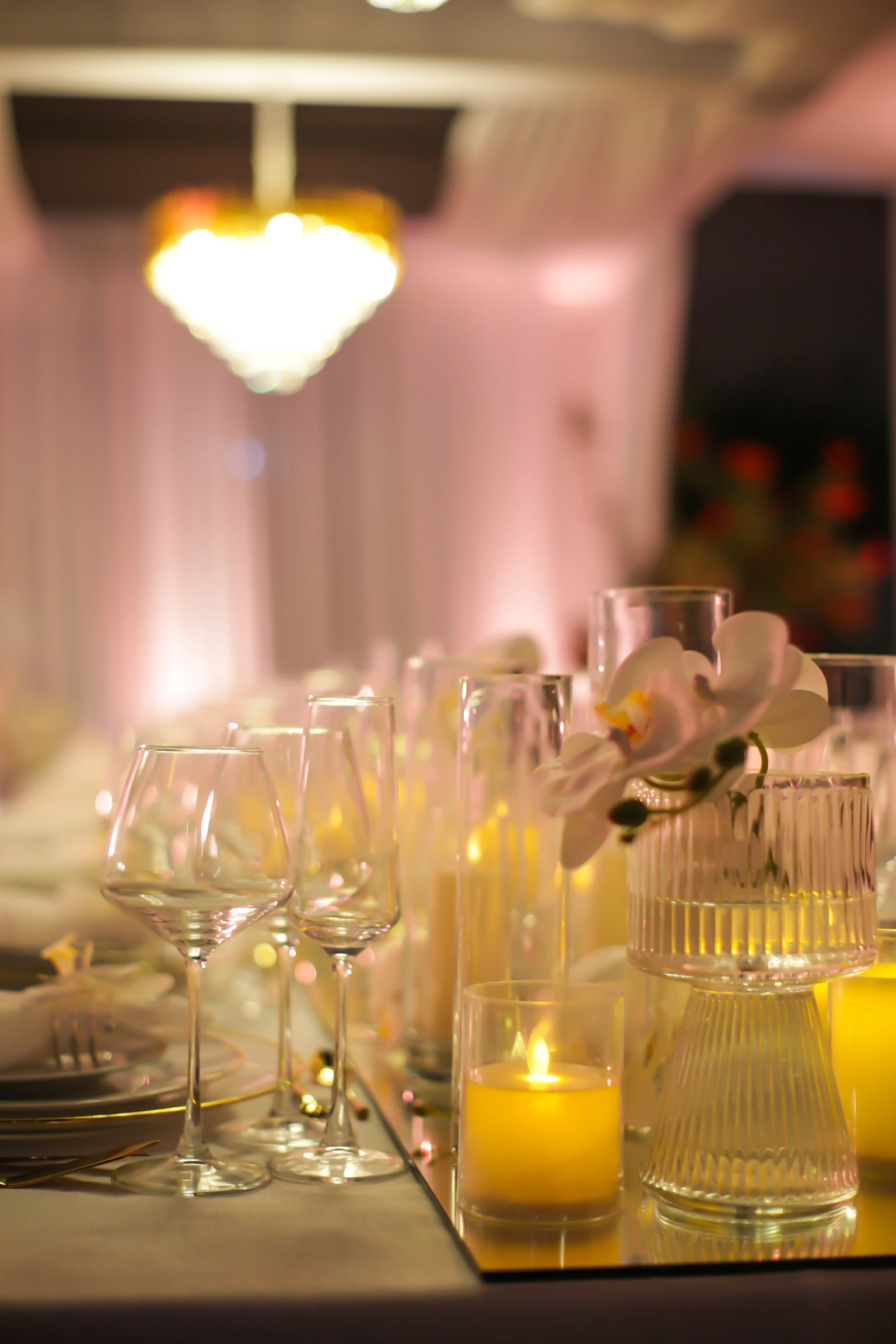 Taças, velas e um vaso canelado com orquídea sobre o friso de espelho, com um lustre dourado desfocado ao fundo