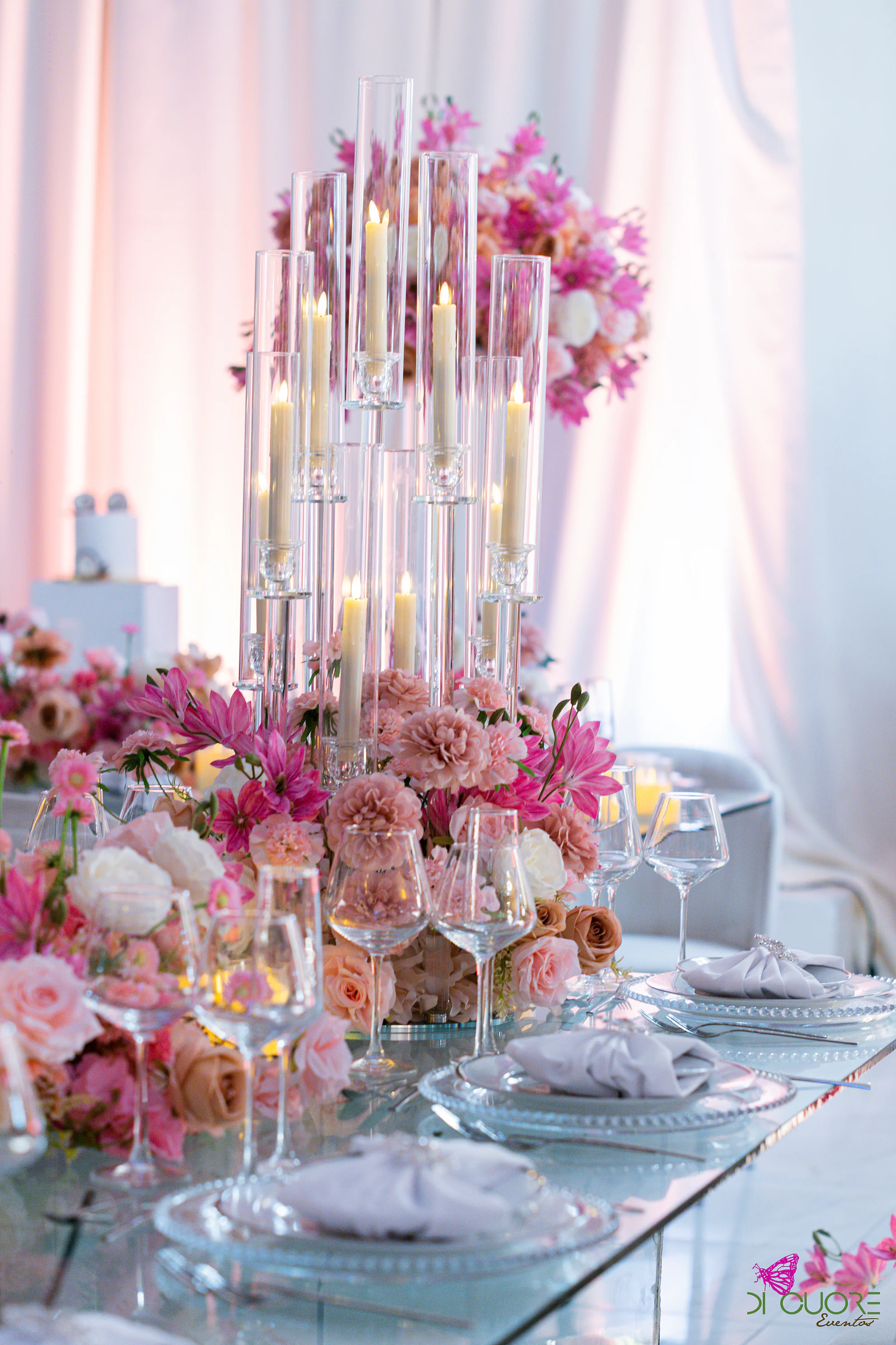 Candelabro de vidro com velas altas entre rosas e flores cor-de-rosa, sobre uma mesa de vidro posta com guardanapos cinzentos