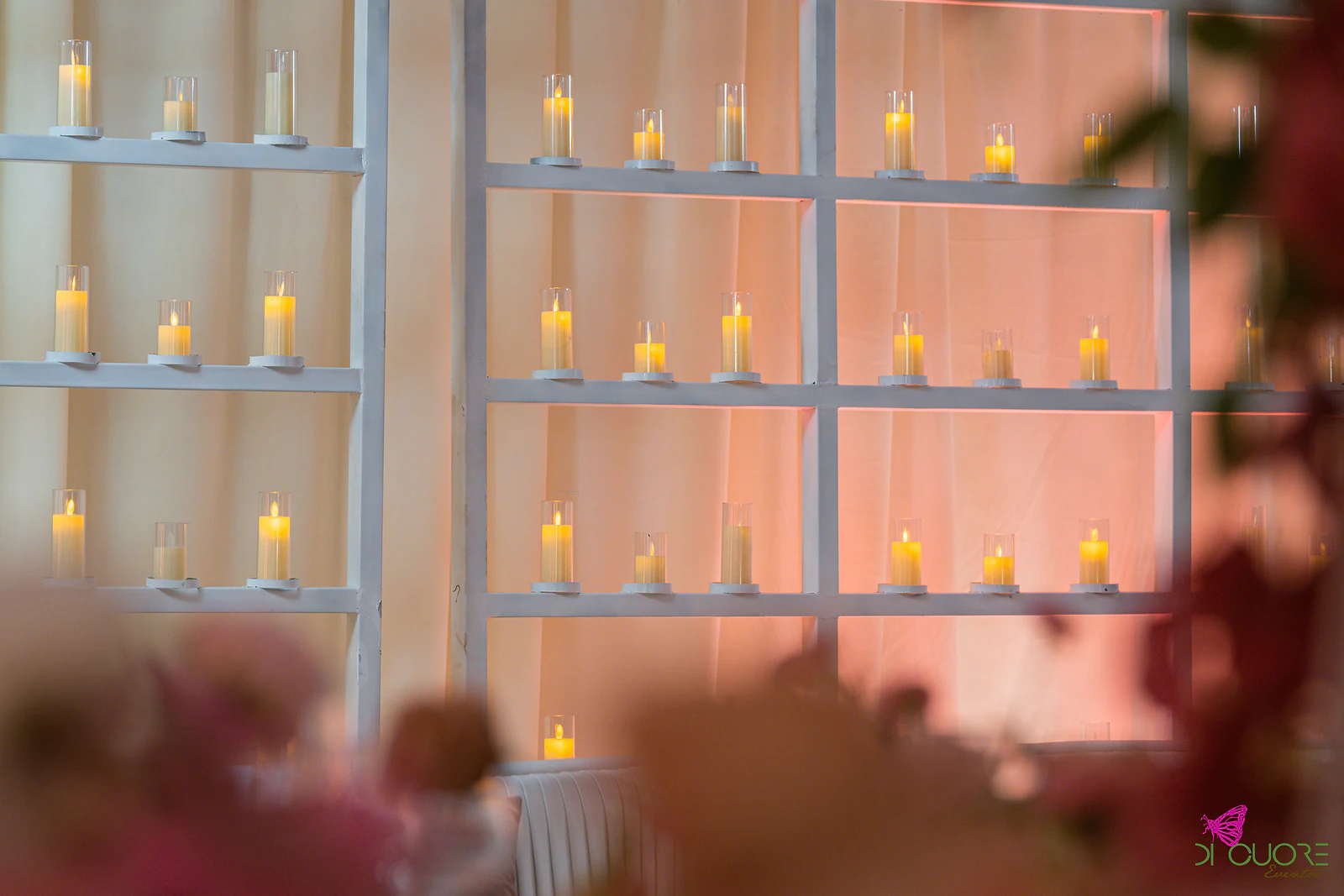 Estantes brancas com velas em cilindros de vidro diante de um cortinado cor de pêssego, com flores desfocadas em primeiro plano