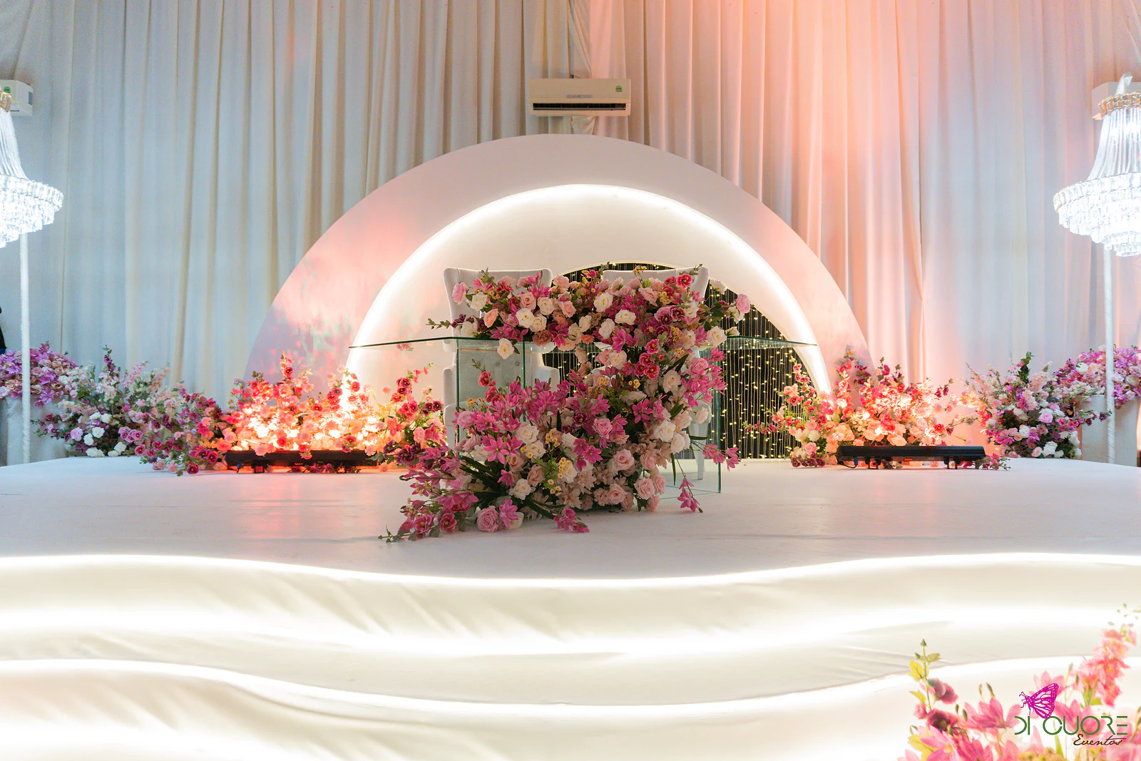Palco com arco branco de contorno luminoso, mesa de vidro coberta de flores em cascata e lustres de cristal de cada lado