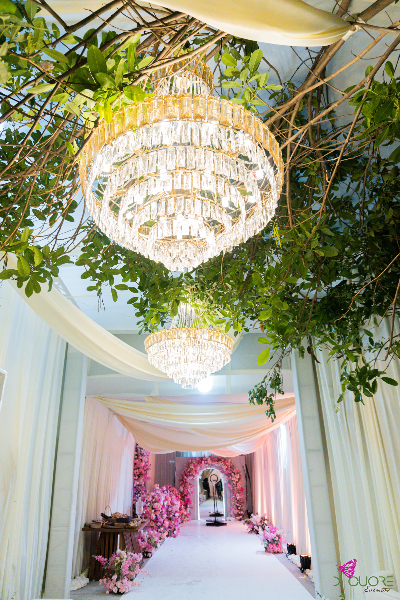Lustres de cristal pendurados de ramos de árvore e panos creme sobre um corredor branco, com flores cor-de-rosa ao fundo