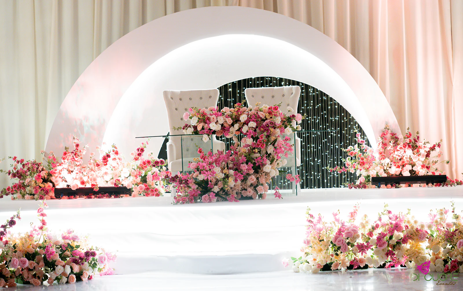 Mesa dos noivos com duas poltronas brancas e arranjos de flores cor-de-rosa, diante de um arco branco e de uma cortina de luzes, sobre um estrado branco