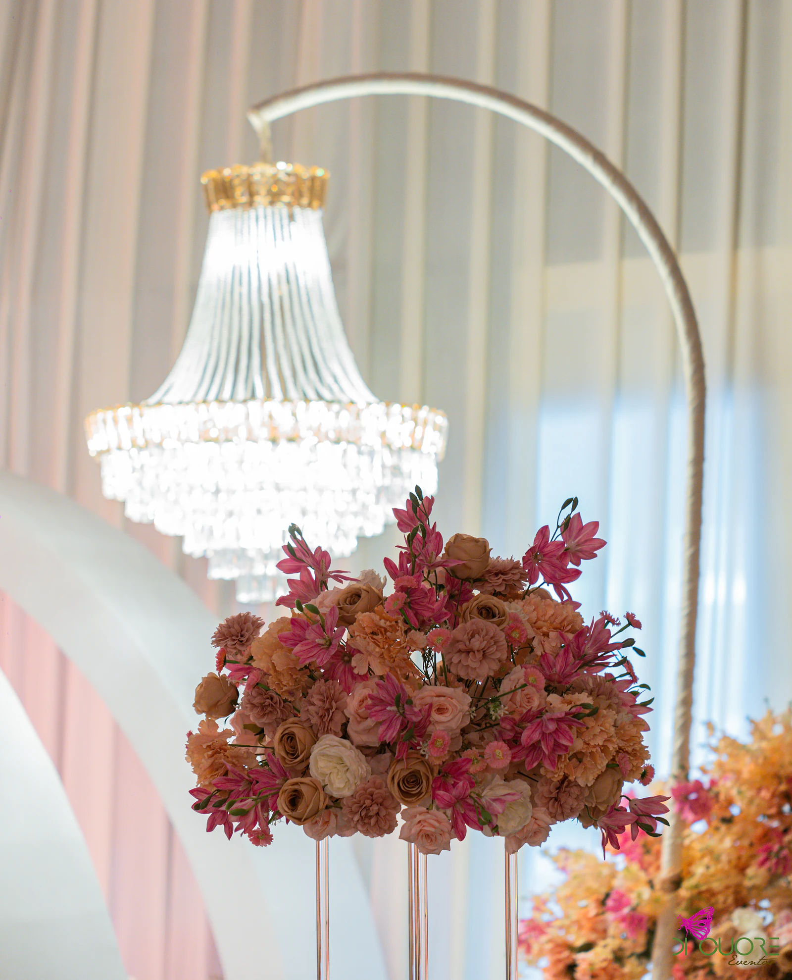 Arranjo alto de rosas e flores cor-de-rosa e coral sobre suportes transparentes, com um lustre de cristal ao fundo
