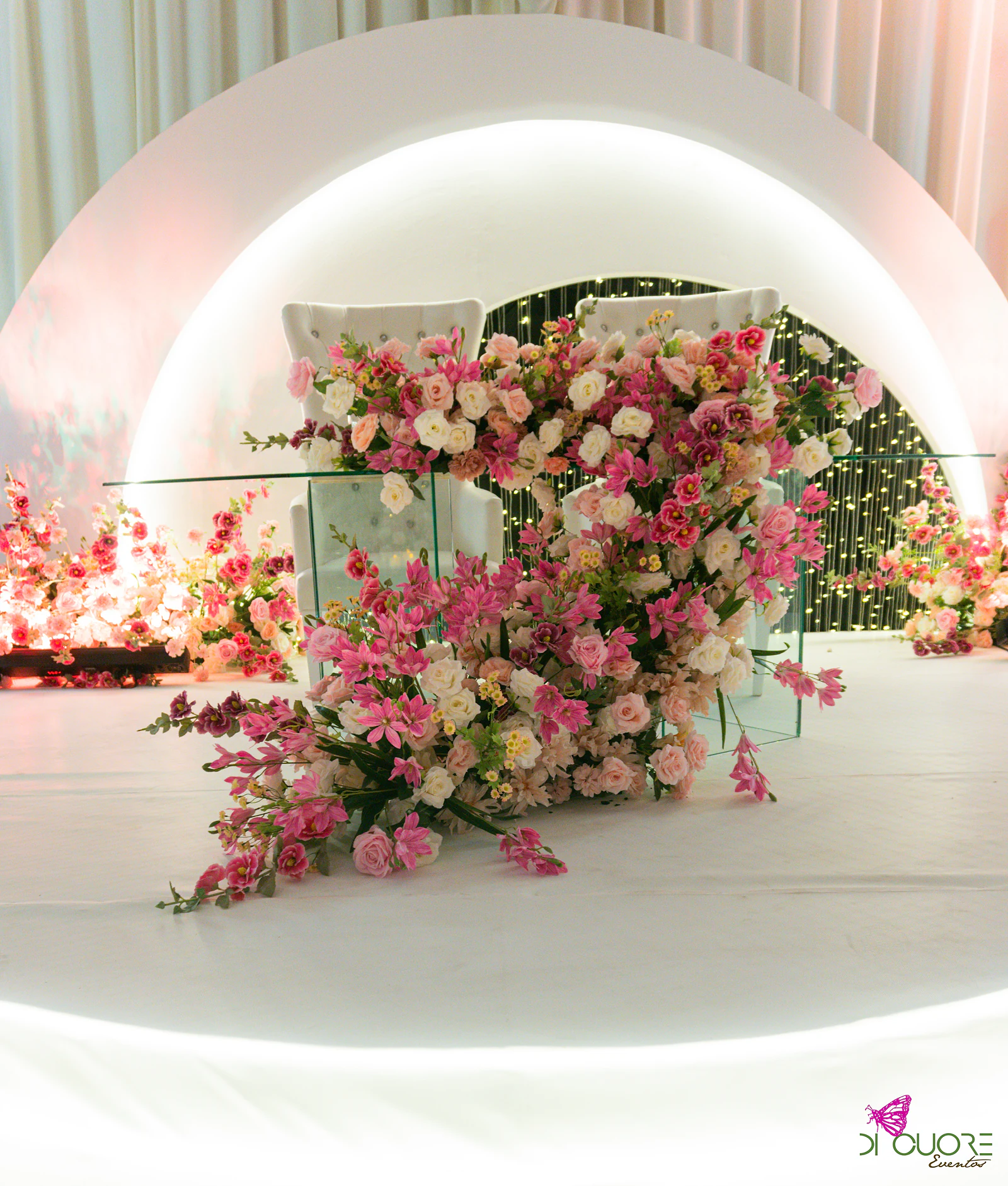 Cadeirões brancos sob um arco de luz, atrás de uma mesa de vidro coberta de rosas e flores cor-de-rosa em cascata
