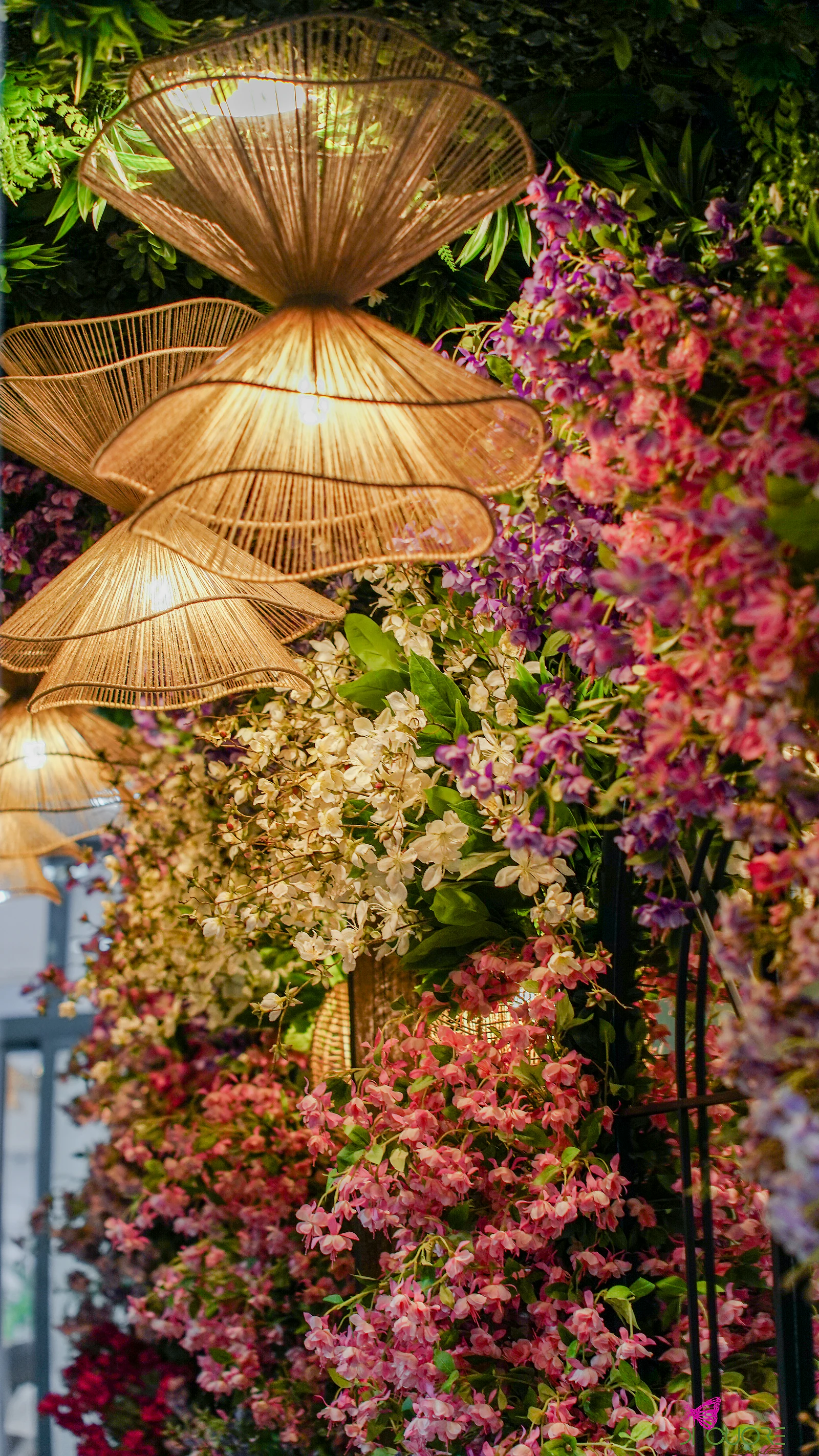 Candeeiros de vime suspensos sobre uma parede de flores cor-de-rosa, brancas e lilás