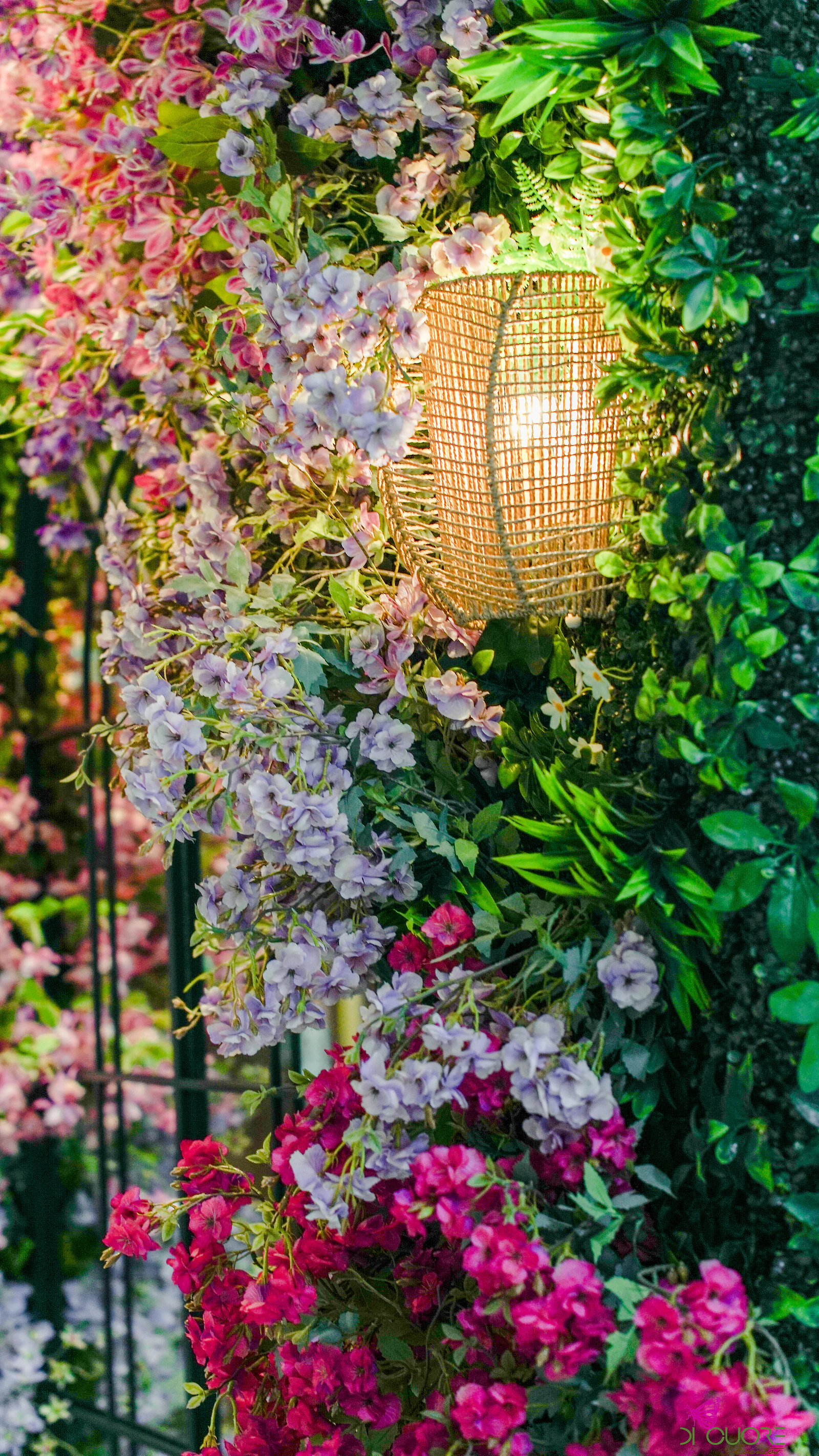 Parede de flores lilás pendentes e fúcsia com um candeeiro de vime aceso, junto a folhagem verde