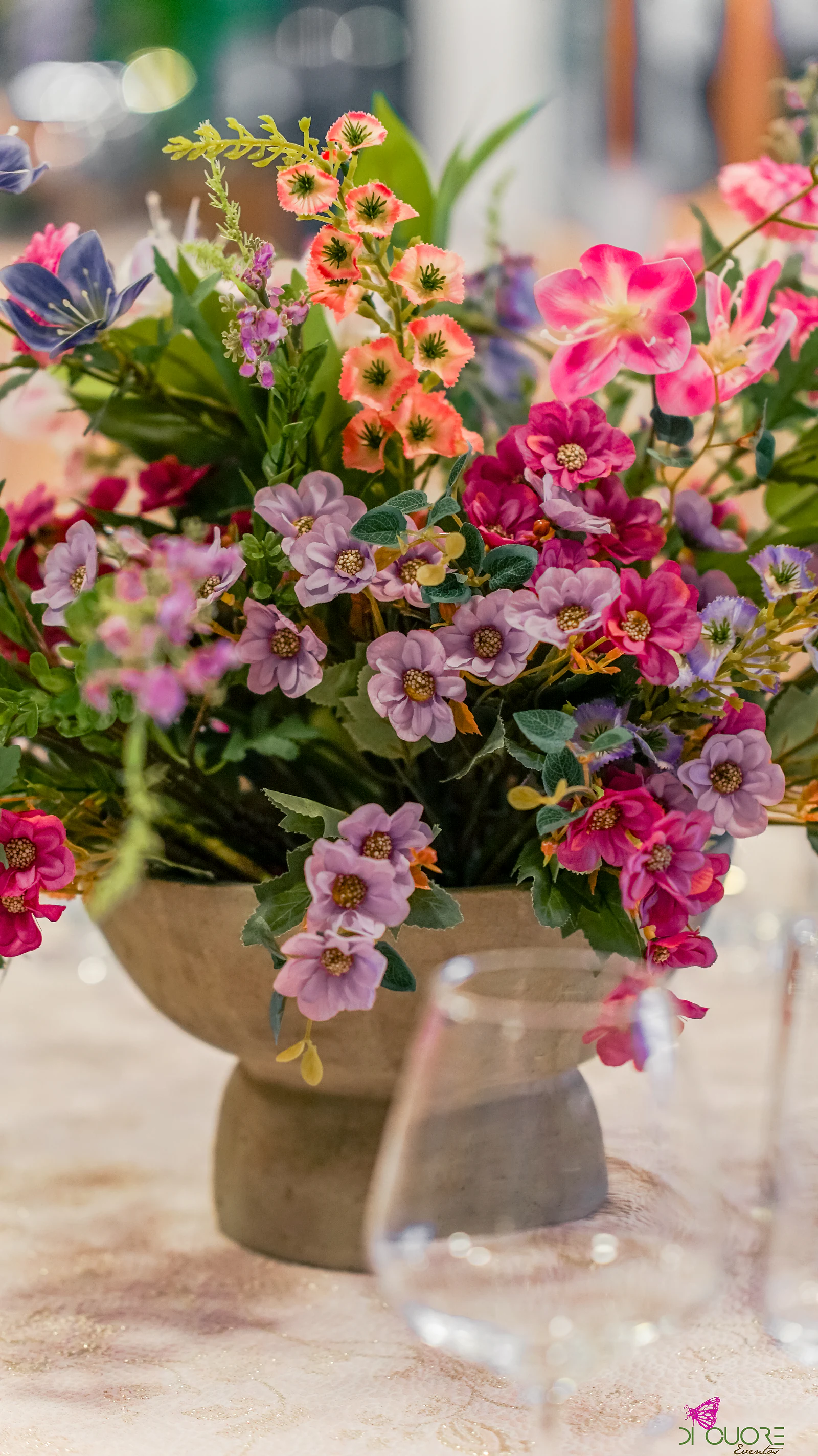 Arranjo numa taça de pedra com flores lilás, fúcsia, cor-de-rosa e laranja sobre a toalha bege