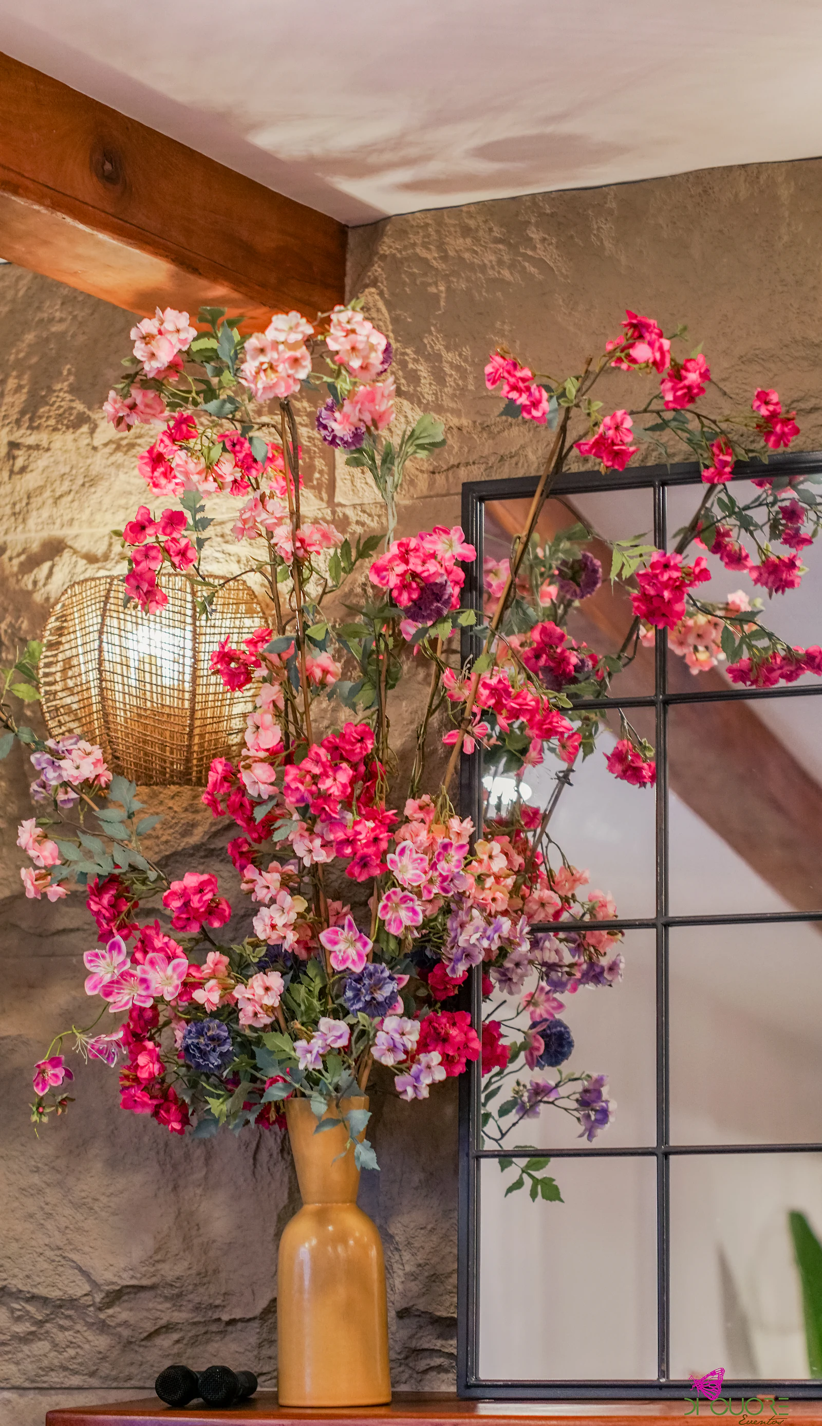 Ramo alto de flores cor-de-rosa e fúcsia num vaso ocre, junto a uma parede de pedra e a um candeeiro de vime