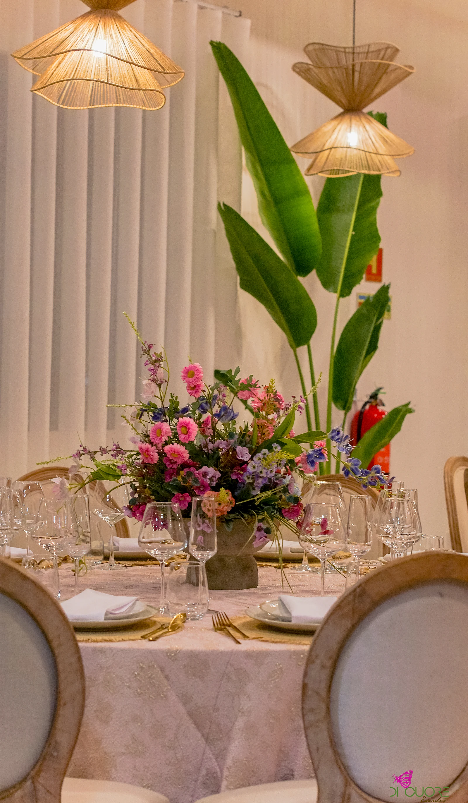 Arranjo de flores cor-de-rosa, azuis e lilás sobre a mesa redonda, com folhas de bananeira e candeeiros de vime diante de cortinas brancas