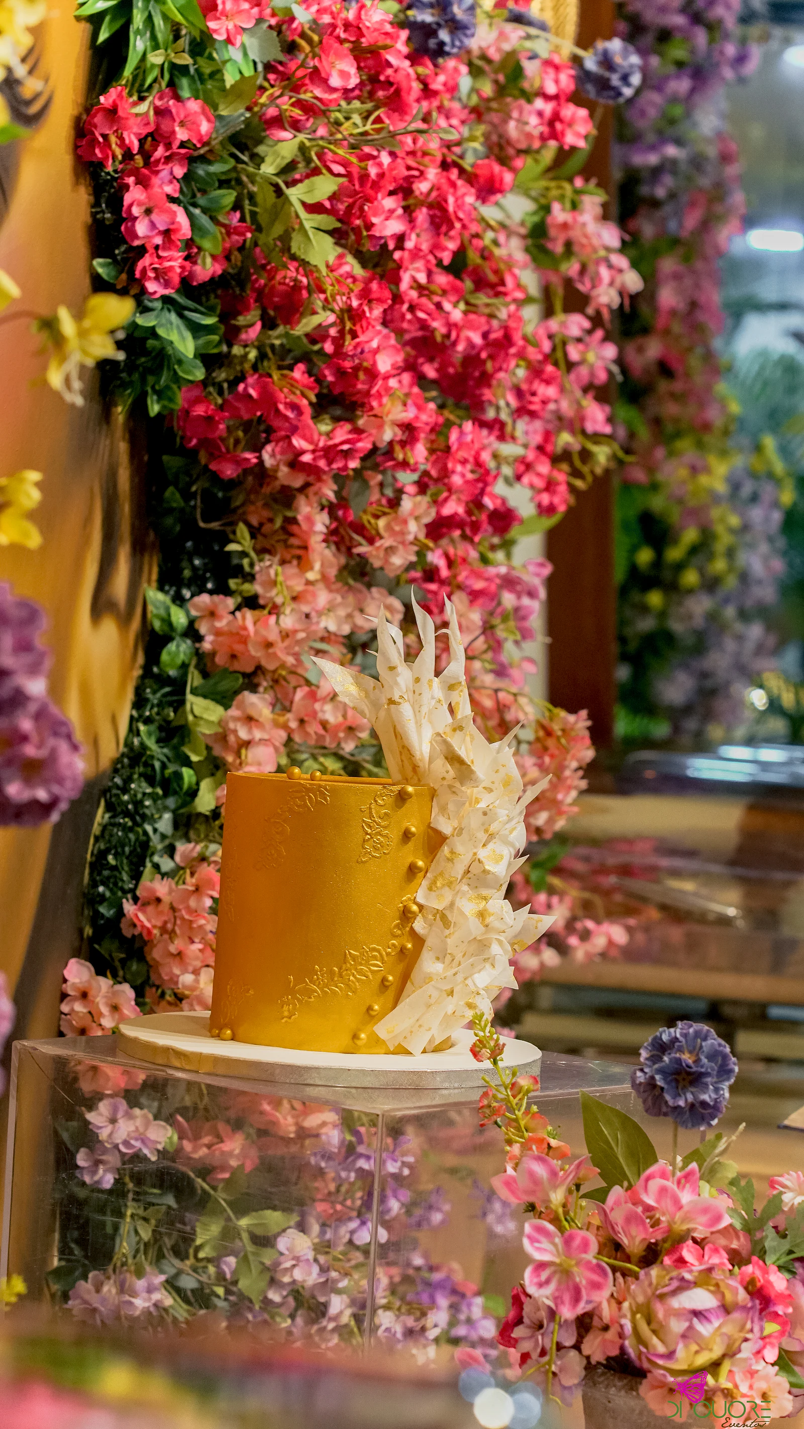 Bolo dourado com lascas brancas sobre um plinto de acrílico, rodeado de flores cor-de-rosa e uma flor lilás