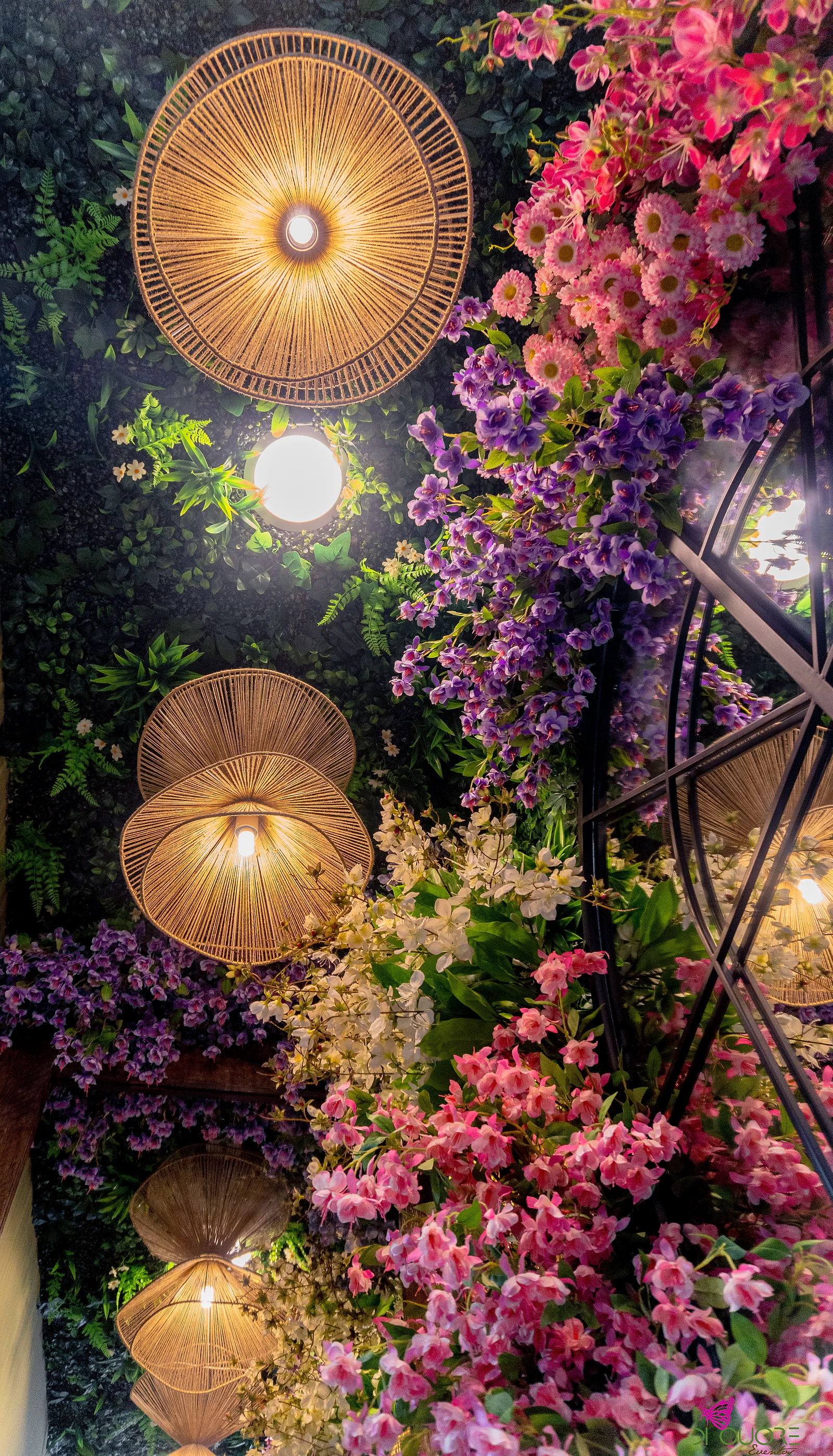 Parede de folhagem escura com candeeiros de vime acesos, entre ramos de flores cor-de-rosa, lilás e brancas