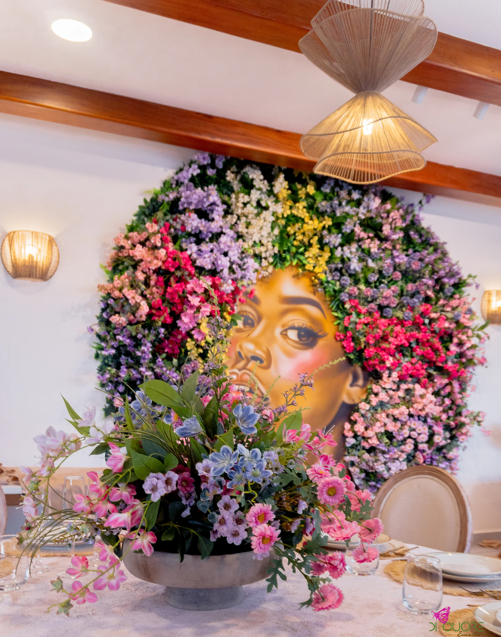 Round mural of a woman’s face framed by pink, lilac and yellow flowers, with a wild-flower arrangement in the foreground and a wicker lamp above