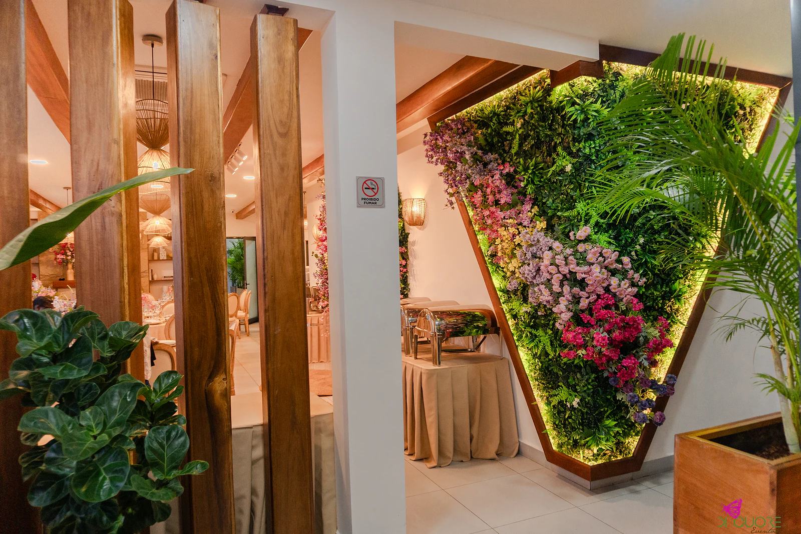 Parede em V de folhagem com flores cor-de-rosa e lilás e contorno de luz quente, ao fundo de um corredor de vigas de madeira