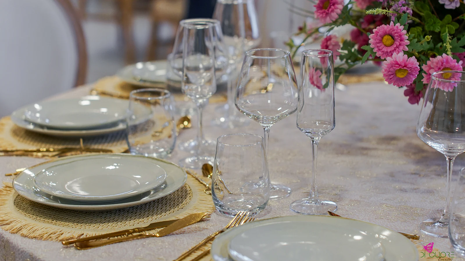 Mesa posta com sousplats de fibra de franja dourada, pratos brancos, talheres dourados, copos e flores cor-de-rosa ao canto