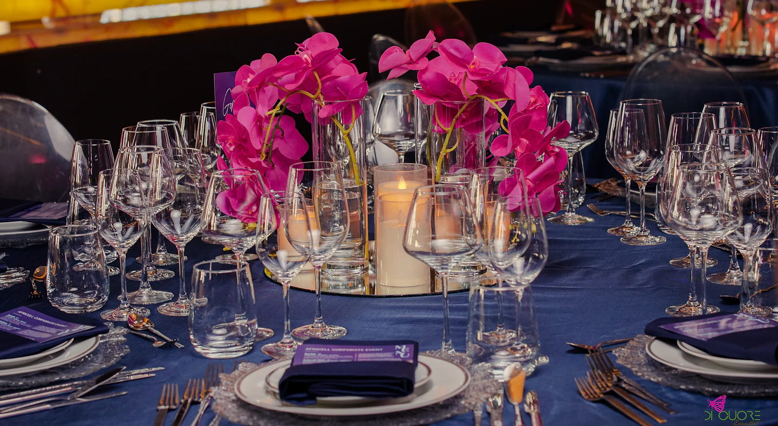 Centrepiece of fuchsia orchids with a candle on a mirrored base, among glasses and dark napkins with the menu, on a blue cloth