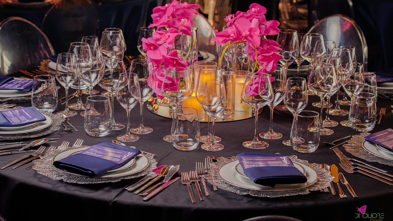 Table with a black cloth laid with textured glass chargers, blue napkins with menus and copper-toned cutlery, and a centrepiece of fuchsia orchids with candles