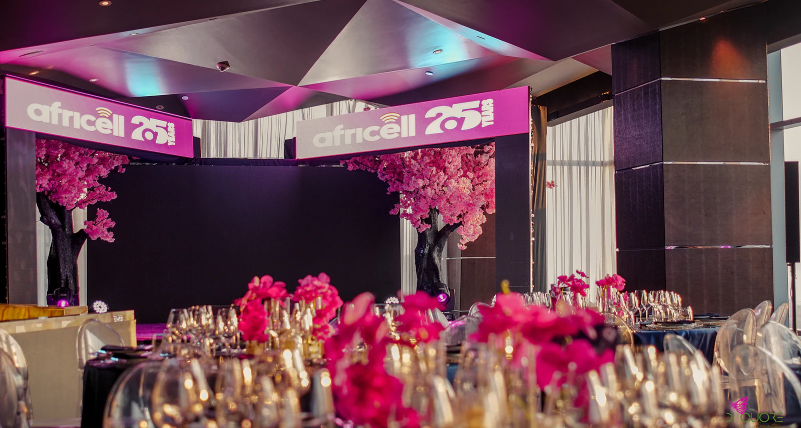 View of the room, with fuchsia orchids and crystal glasses in the foreground and, beyond, the stage with cherry trees in blossom and the arches bearing the brand’s sign
