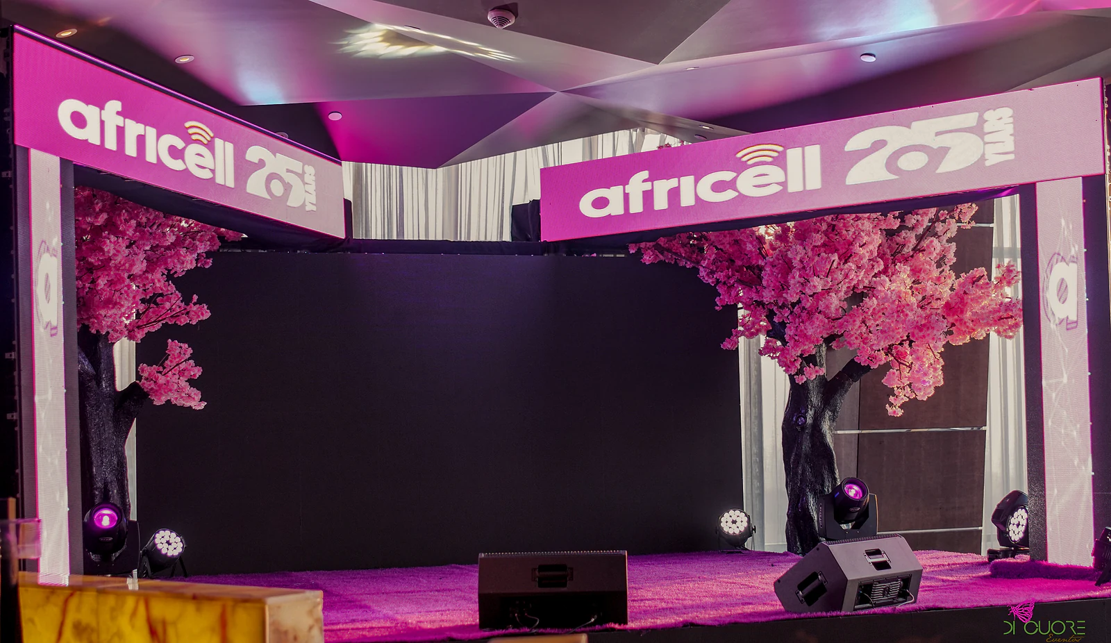 Palco com dois arcos luminosos com o letreiro dos 25 anos da marca, duas cerejeiras em flor e painel escuro ao centro, sob luz magenta
