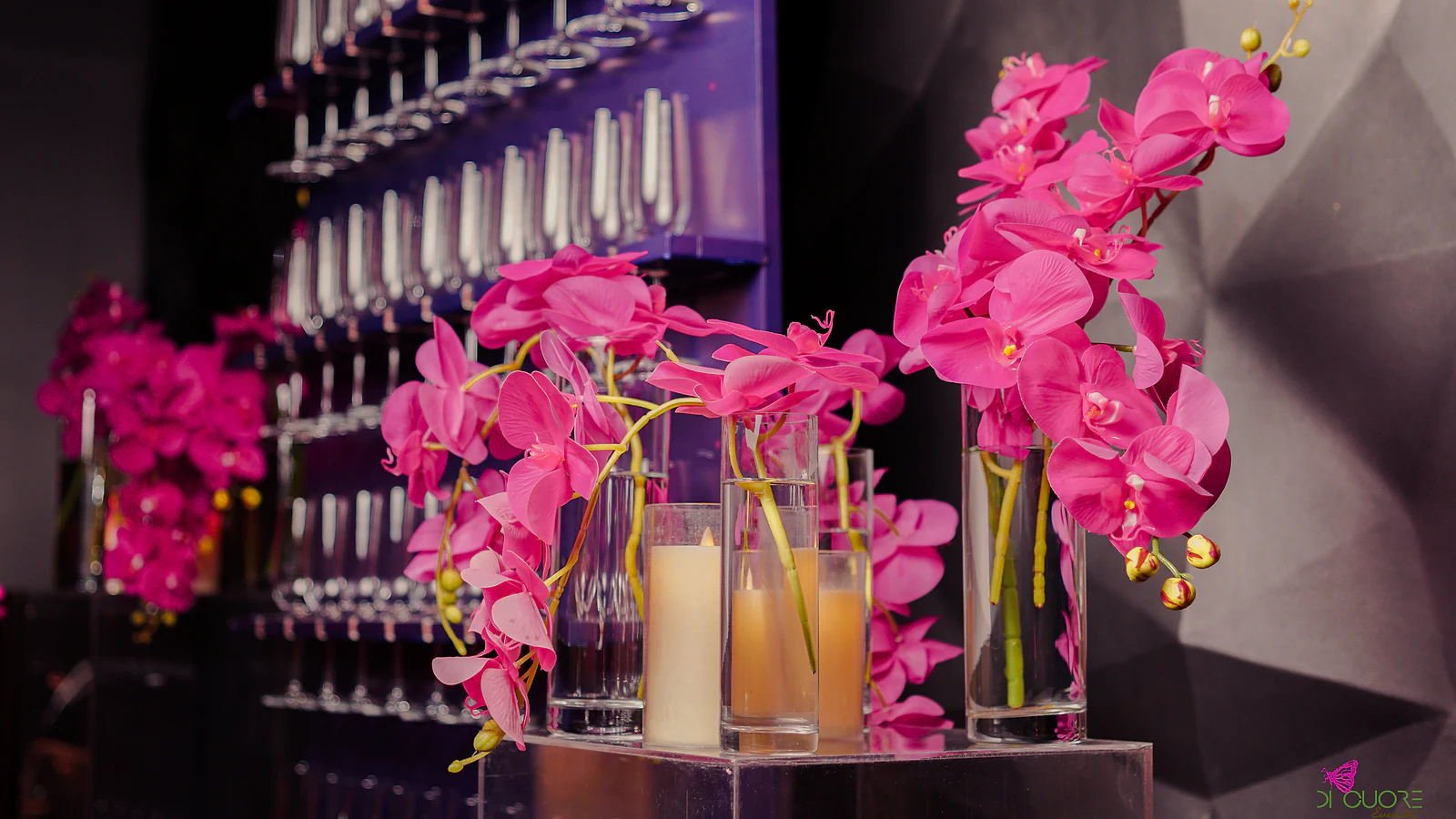 Glass vases of fuchsia orchids and candles on a mirrored plinth, before a wall of glasses in violet light