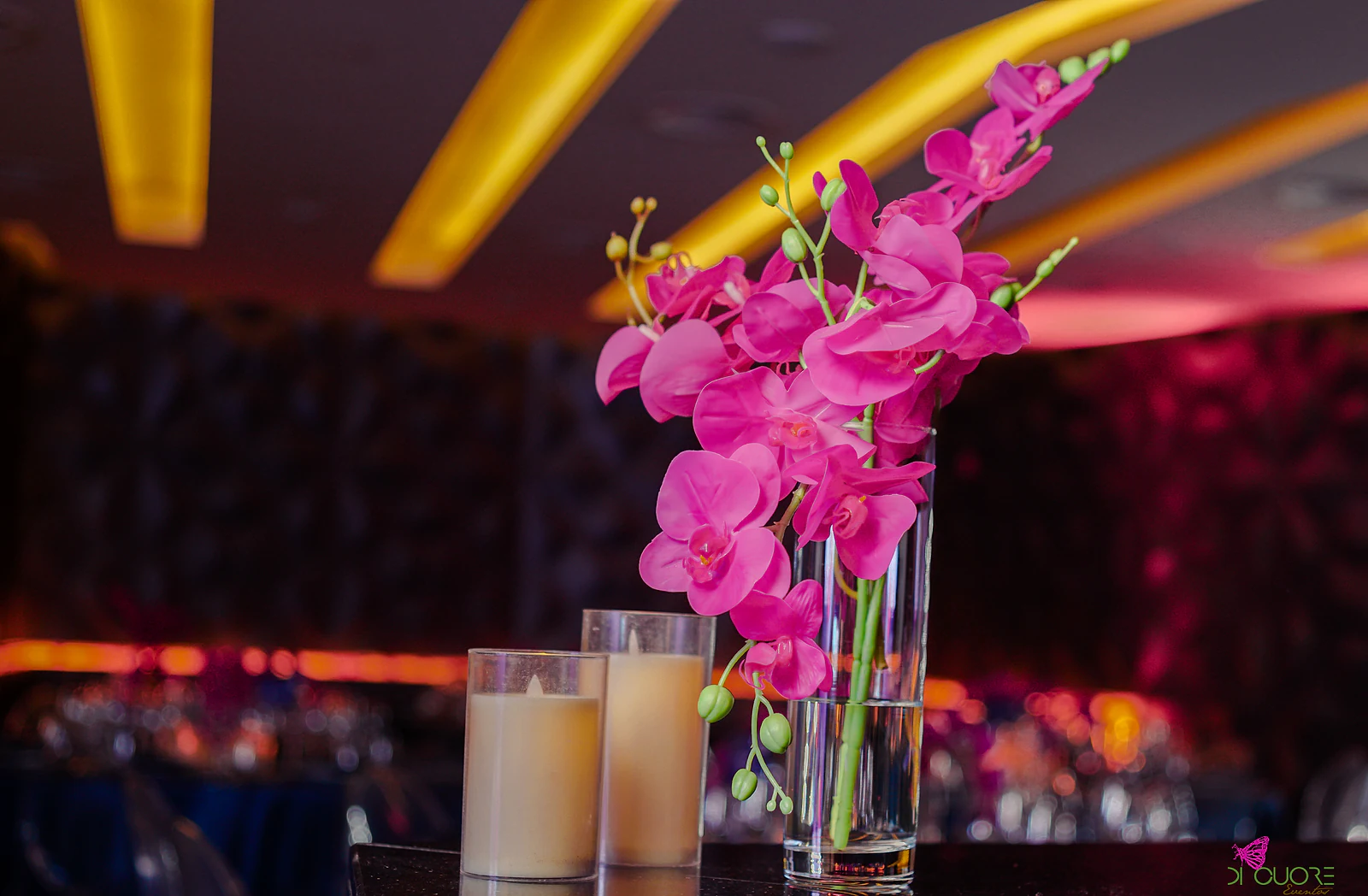 Stem of fuchsia orchids in a glass vase beside candles, with the room in amber light blurred behind