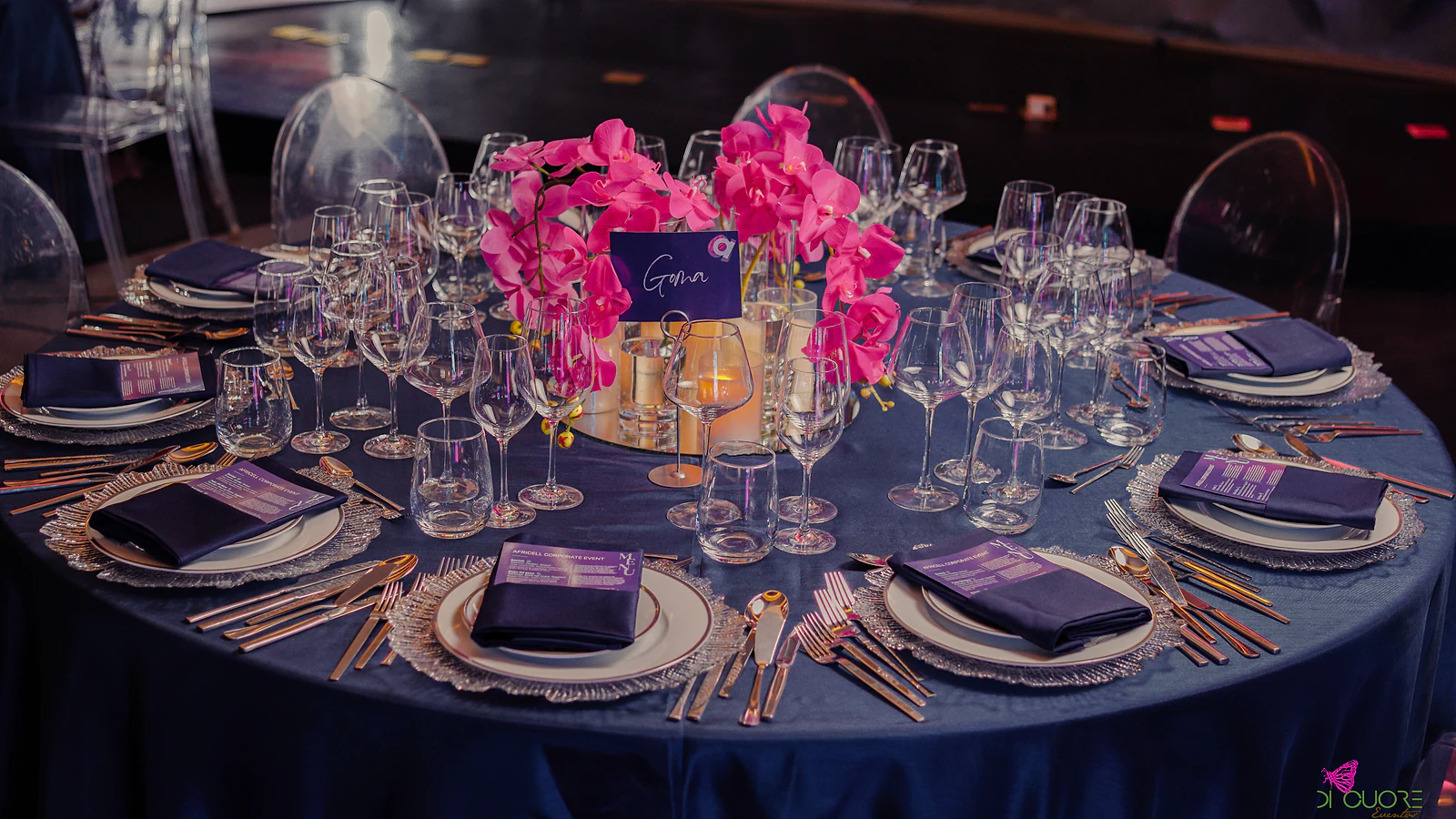 Round table with a blue satin cloth, a table card, fuchsia orchids on a mirror and candles, violet napkins and textured glass chargers