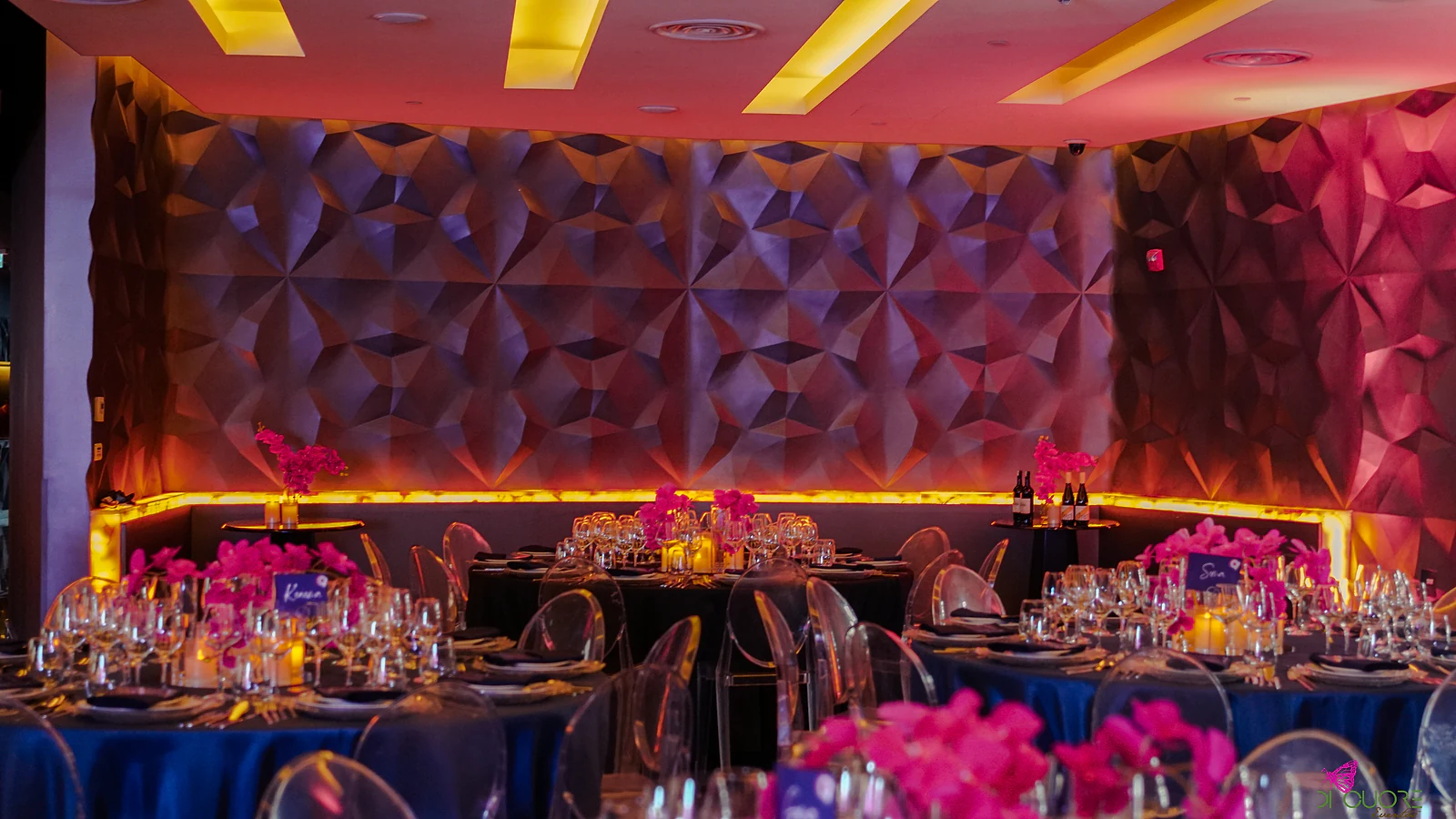 Room with round tables in blue cloths, fuchsia orchids and clear chairs, before the faceted wall lit in orange and fuchsia