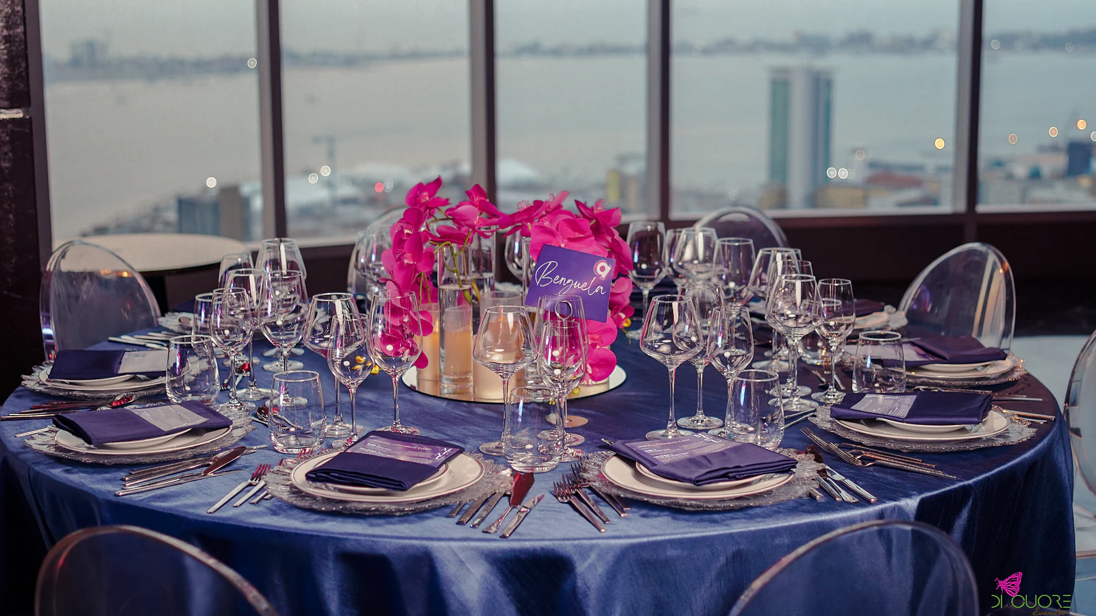 Round table with a blue satin cloth, fuchsia orchids and a table card, before windows over the water and the city skyline