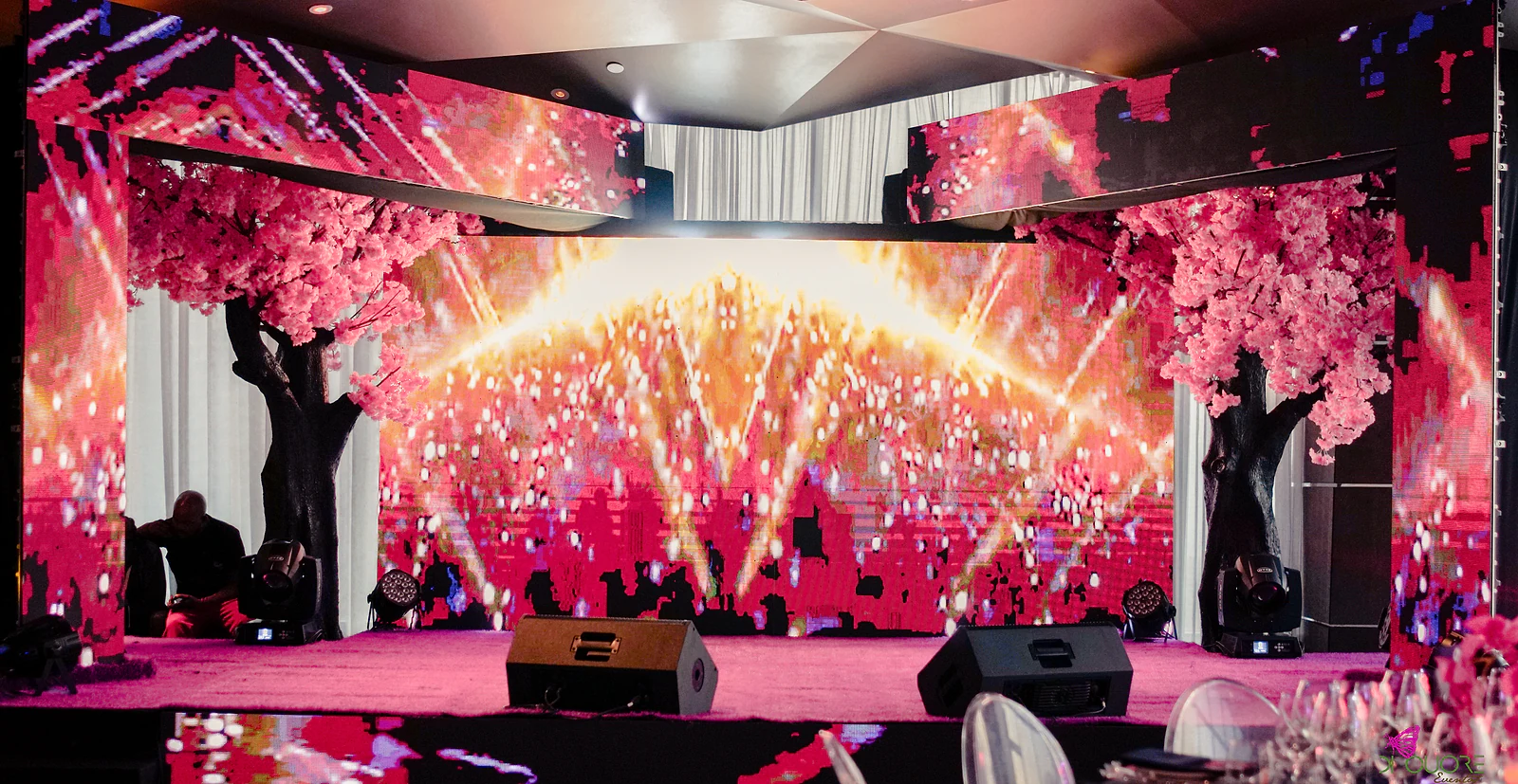 Front view of the stage, with a large screen of red and pink lights between two cherry trees in blossom, beneath the ceiling of the room