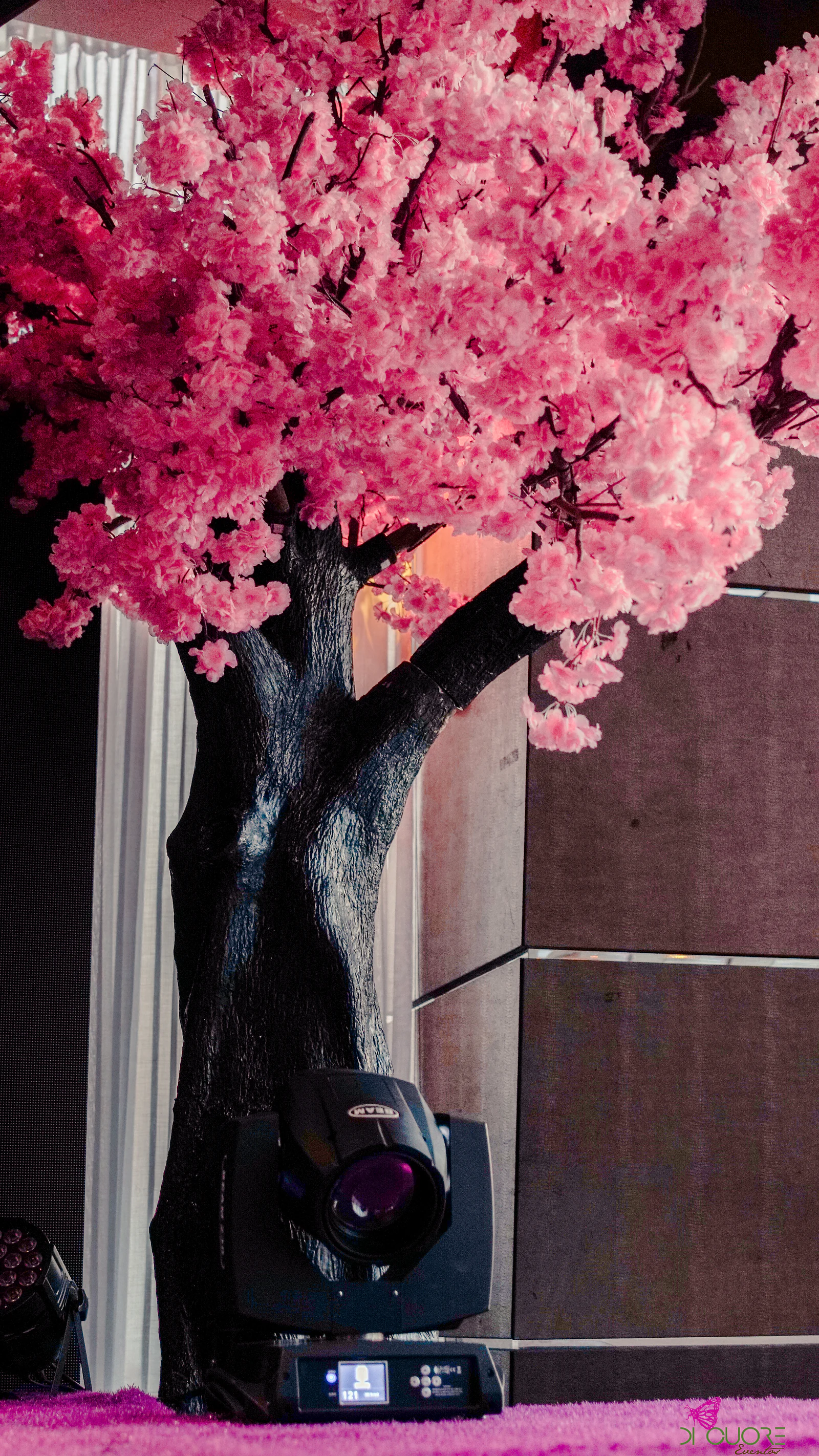Crown of a pink cherry tree in blossom above a dark trunk, with a stage light at its foot and wooden panels behind