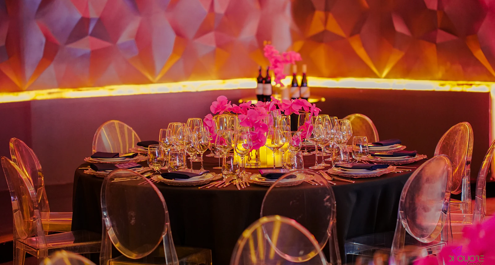 Round table with a black cloth, clear chairs and fuchsia orchids, before the faceted wall with a band of amber light