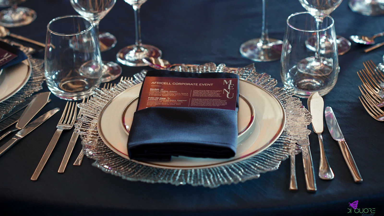 Textured glass charger with a dark napkin and printed menu, between copper-toned cutlery and glasses, on a navy cloth