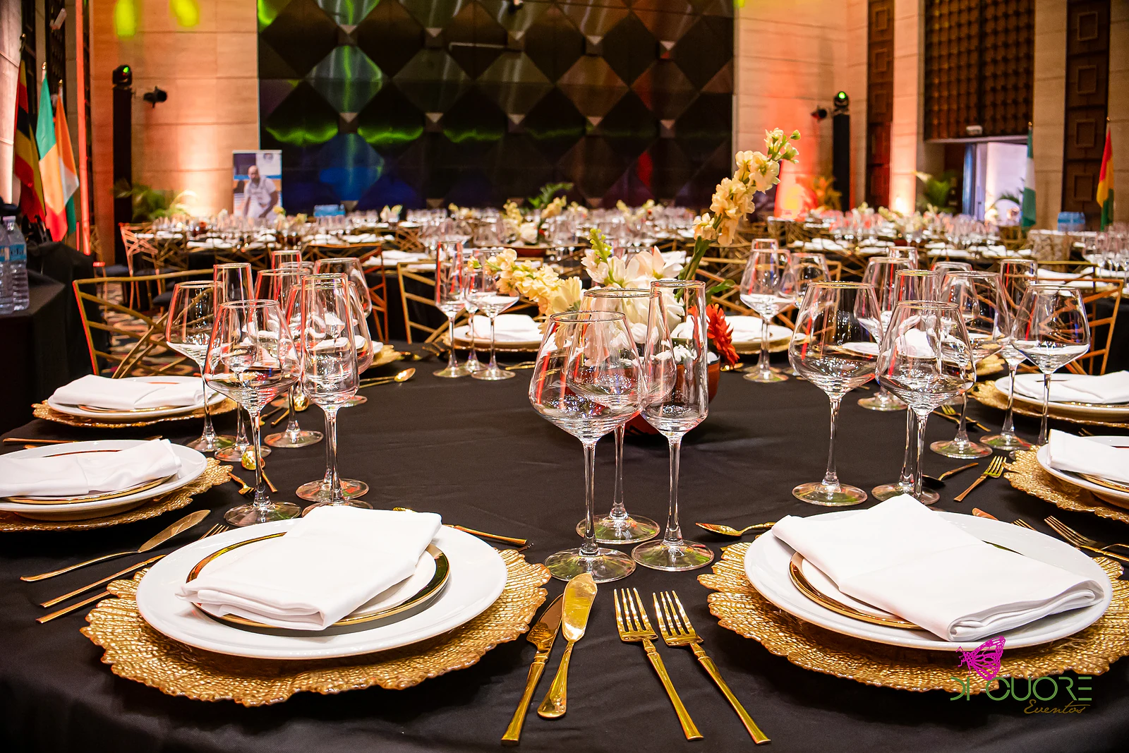 Lugar à mesa com sousplat dourado, prato branco e guardanapo, com copos de cristal e flores ao centro